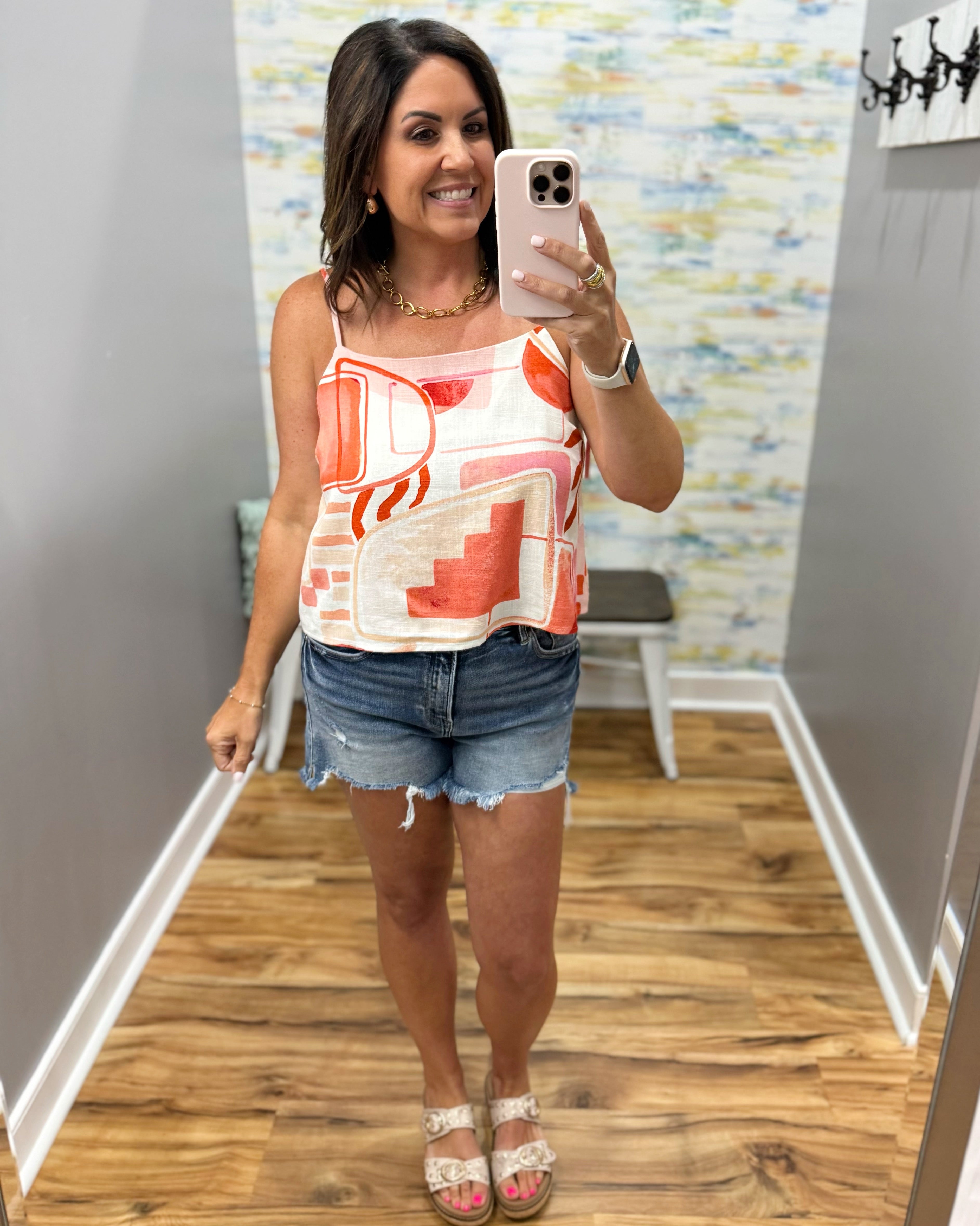 Woman taking a mirror selfie wearing a colorful tank top and denim shorts in a room with patterned wallpaper.