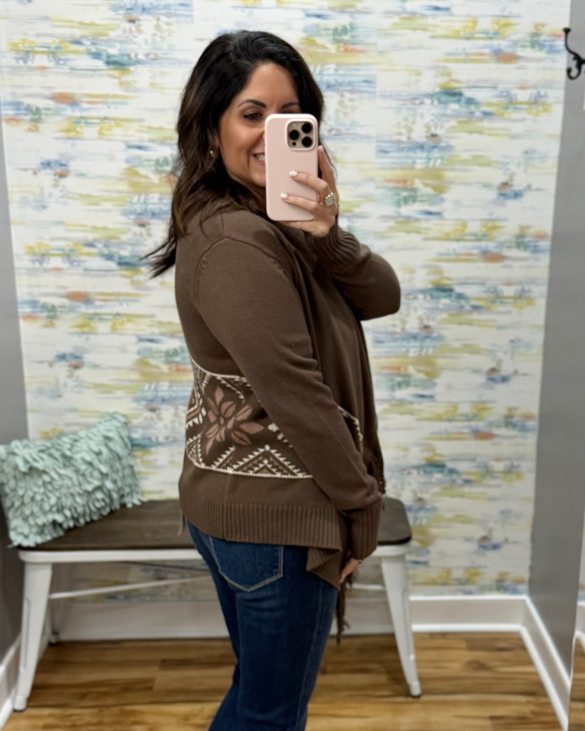 A woman takes a mirror selfie wearing a brown knit cardigan with a geometric patterned panel on the back, paired with blue jeans in a room with patterned wallpaper.