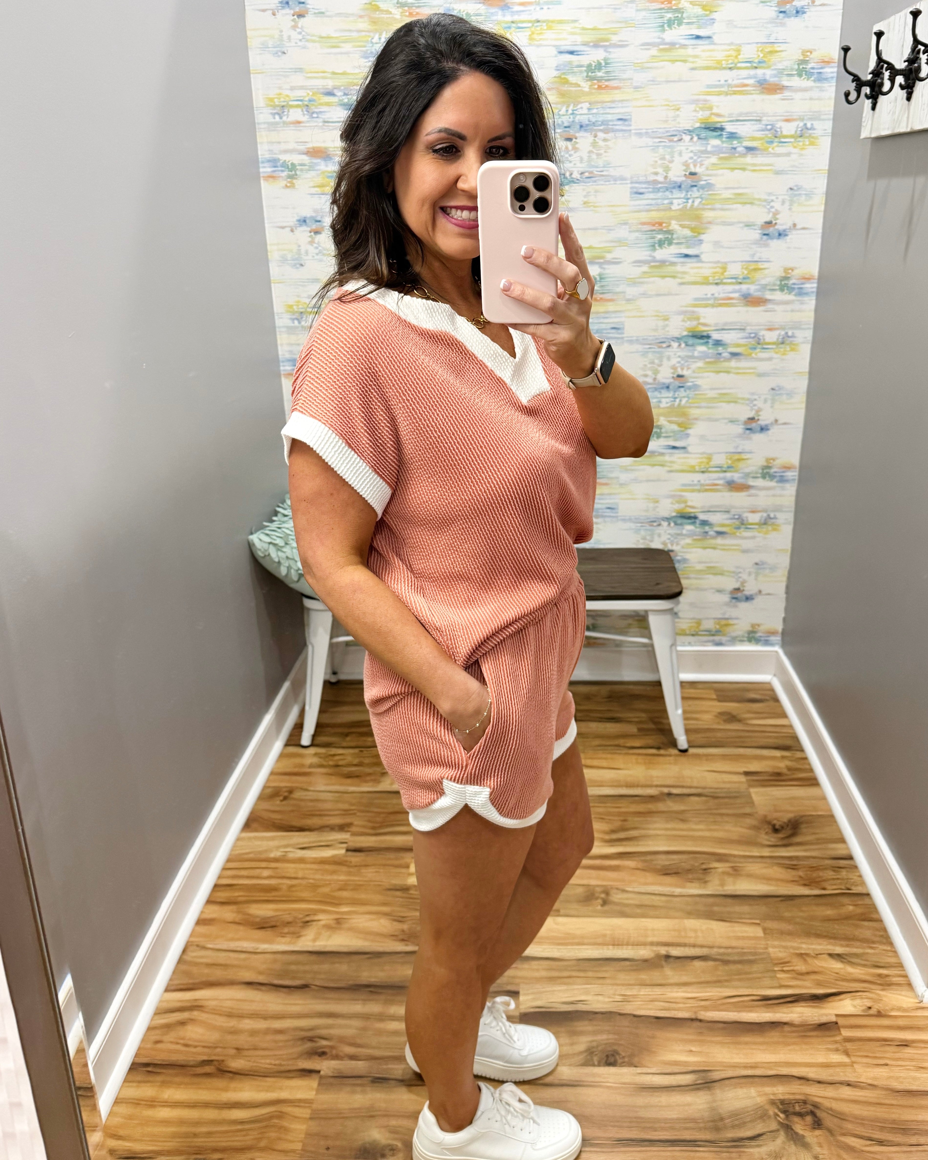 Woman taking a mirror selfie wearing a pink outfit in a room with wooden flooring and a colorful wall.