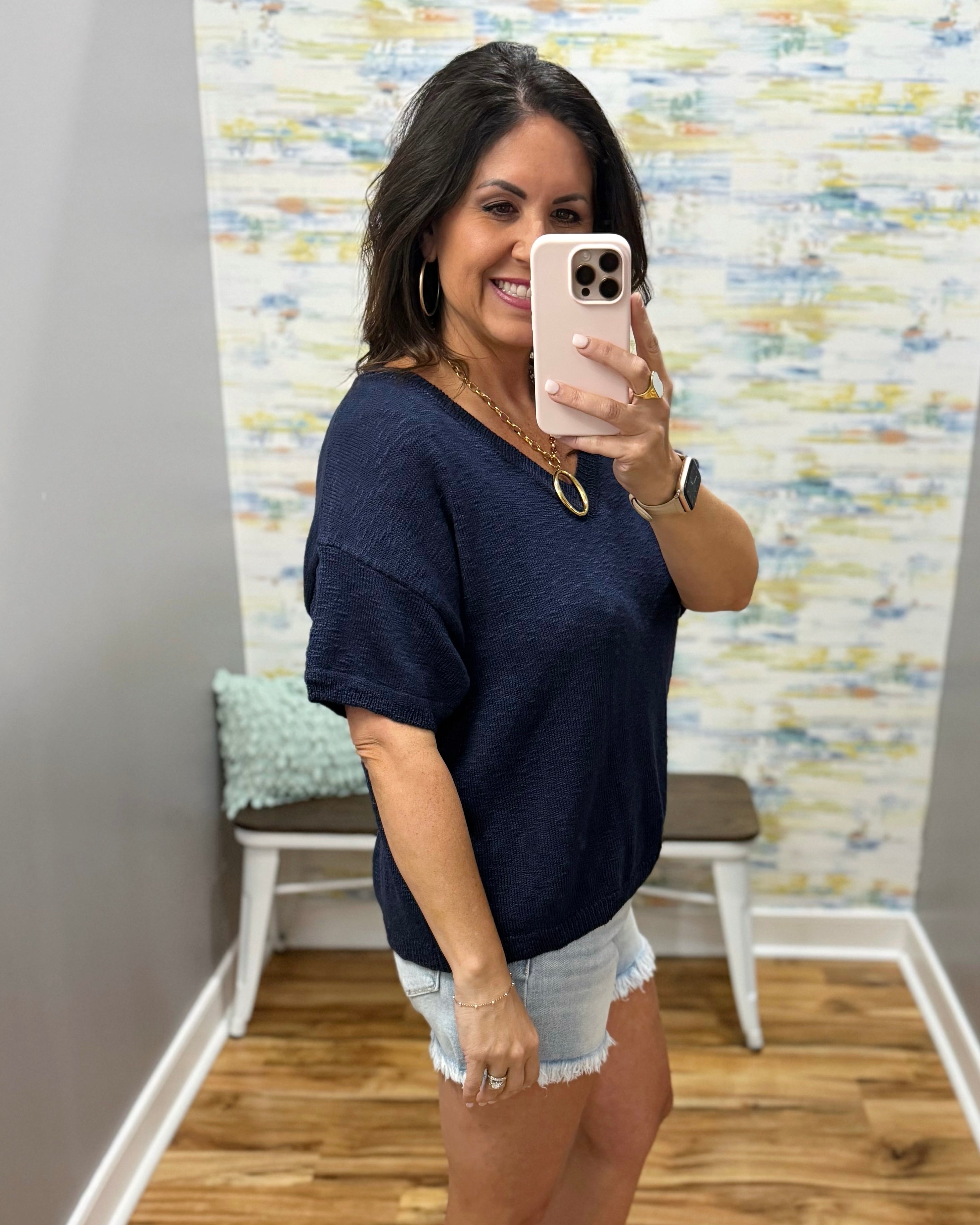 Woman taking a mirror selfie wearing a navy blue shirt and denim shorts in a room with patterned wallpaper.