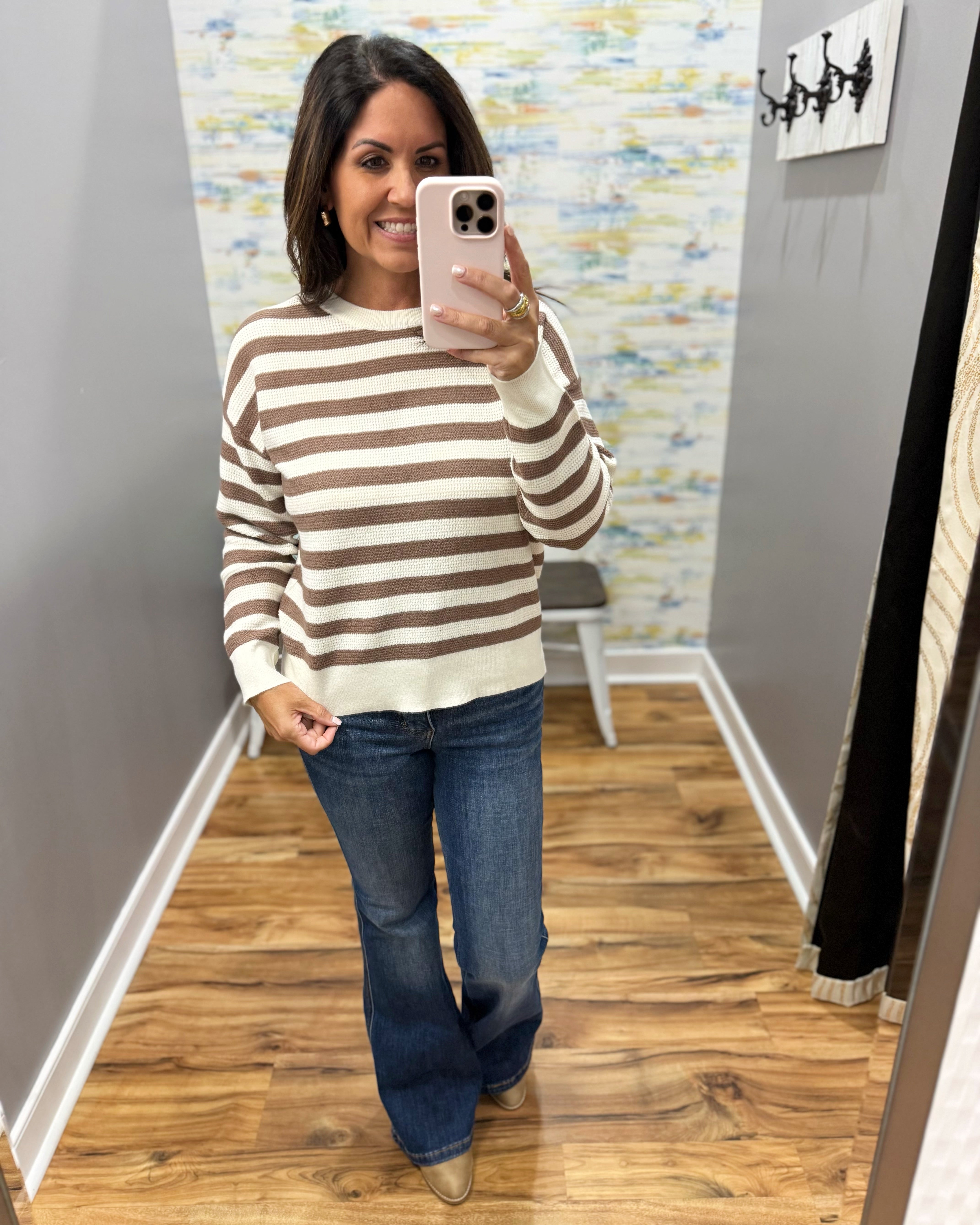 A woman takes a mirror selfie in a fitting room wearing a brown and cream striped crewneck sweater, blue flare jeans, and tan pointed-toe flats.