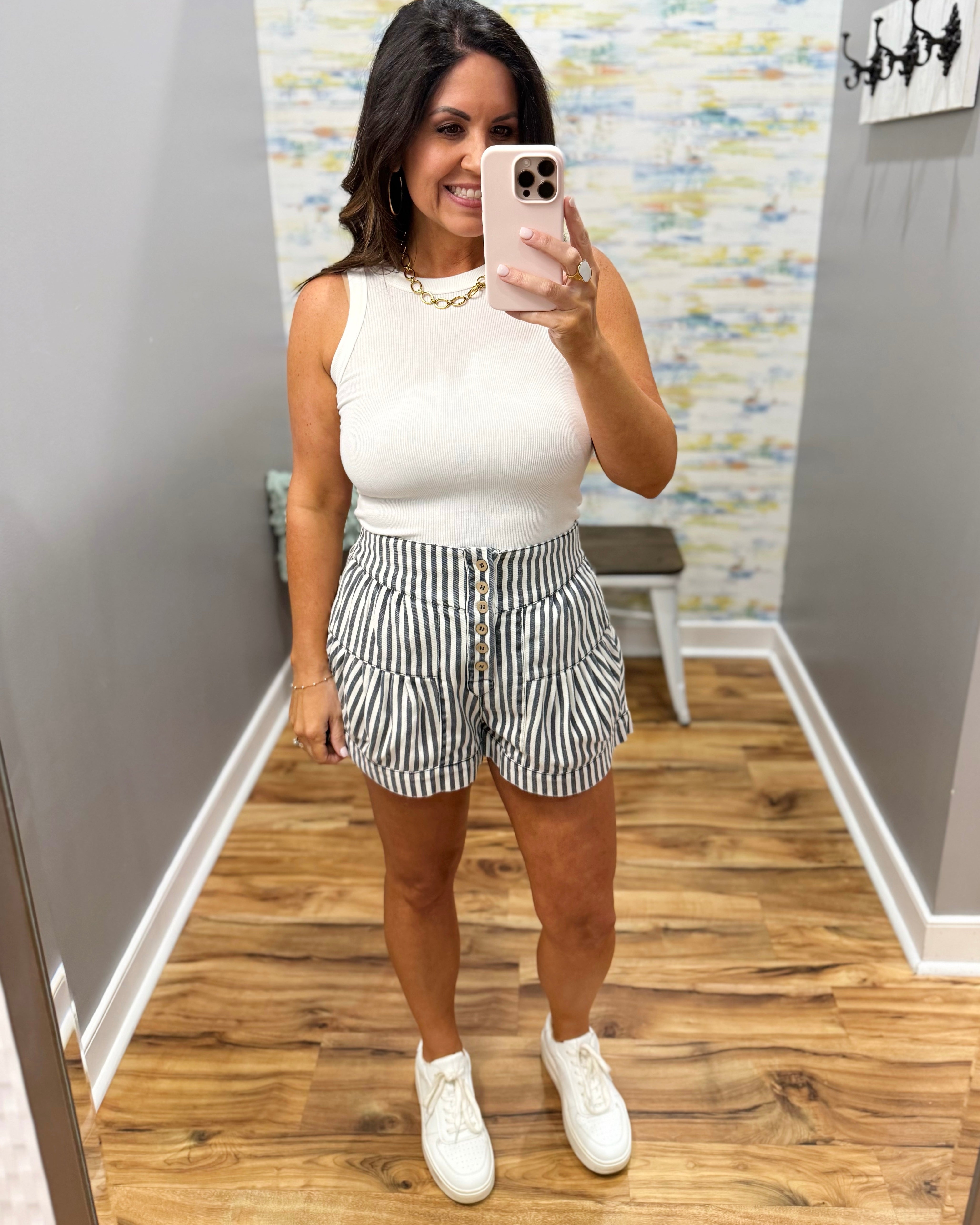 Woman taking a mirror selfie wearing a white sleeveless top and striped shorts.