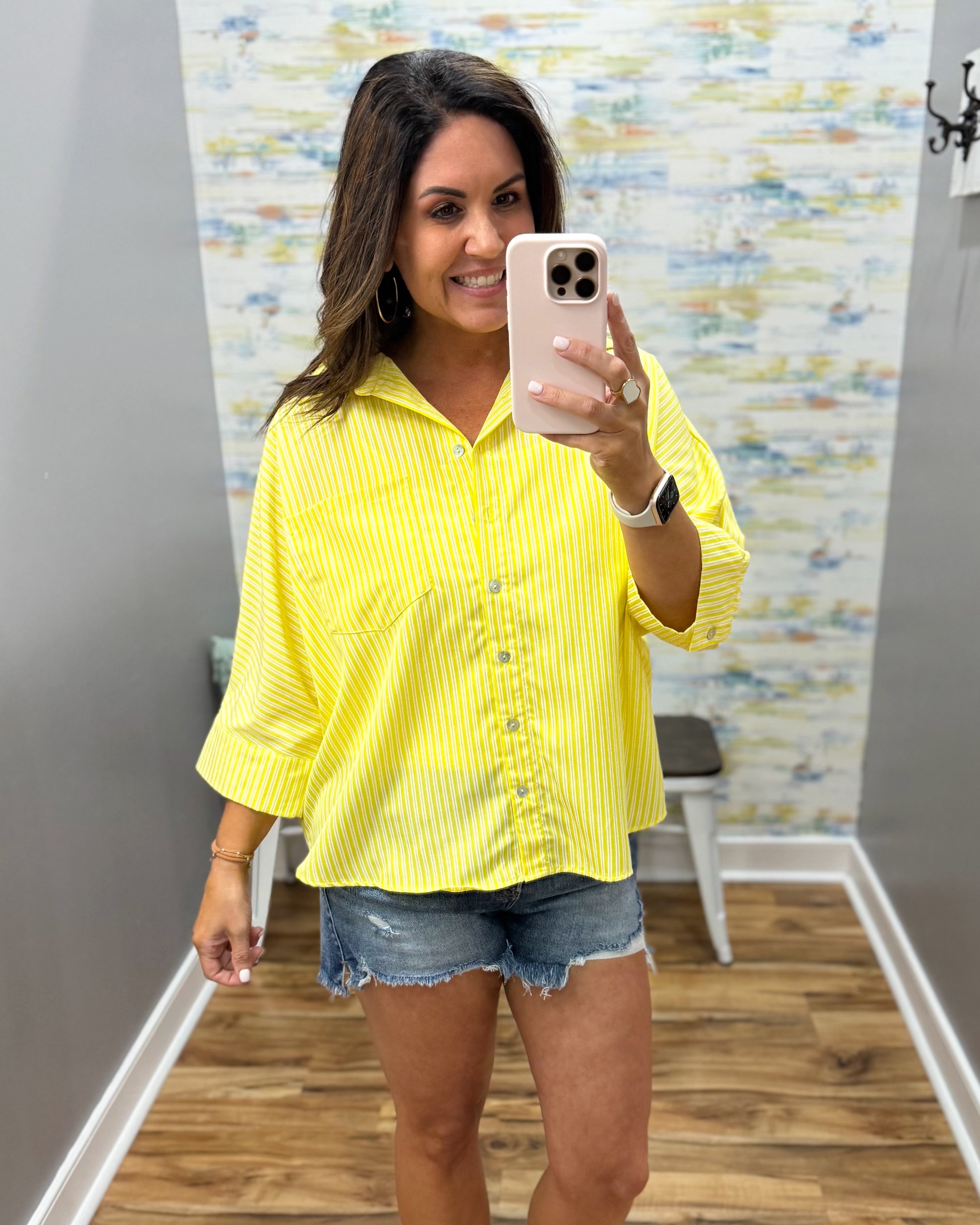Woman taking a mirror selfie wearing a yellow striped shirt and denim shorts.