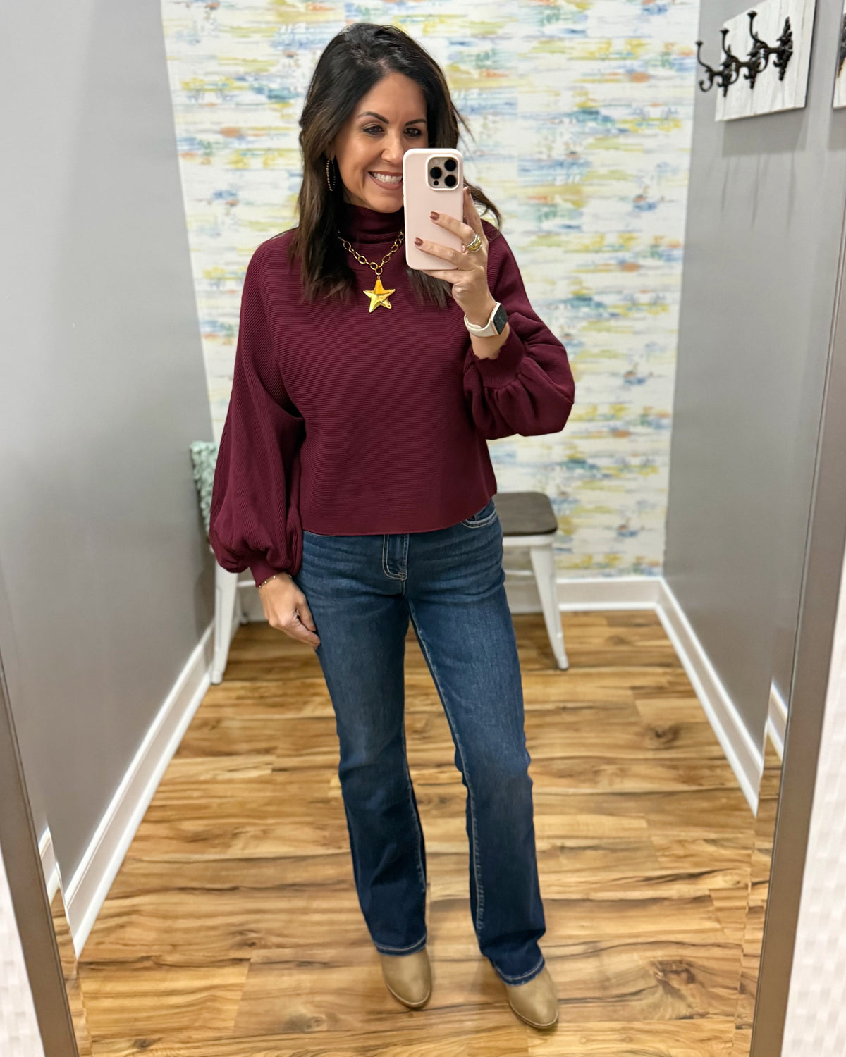 A woman takes a mirror selfie in a fitting room wearing a burgundy long-sleeve mock-neck top with balloon sleeves, mica bootcut jeans, and corkys beige ankle boots.