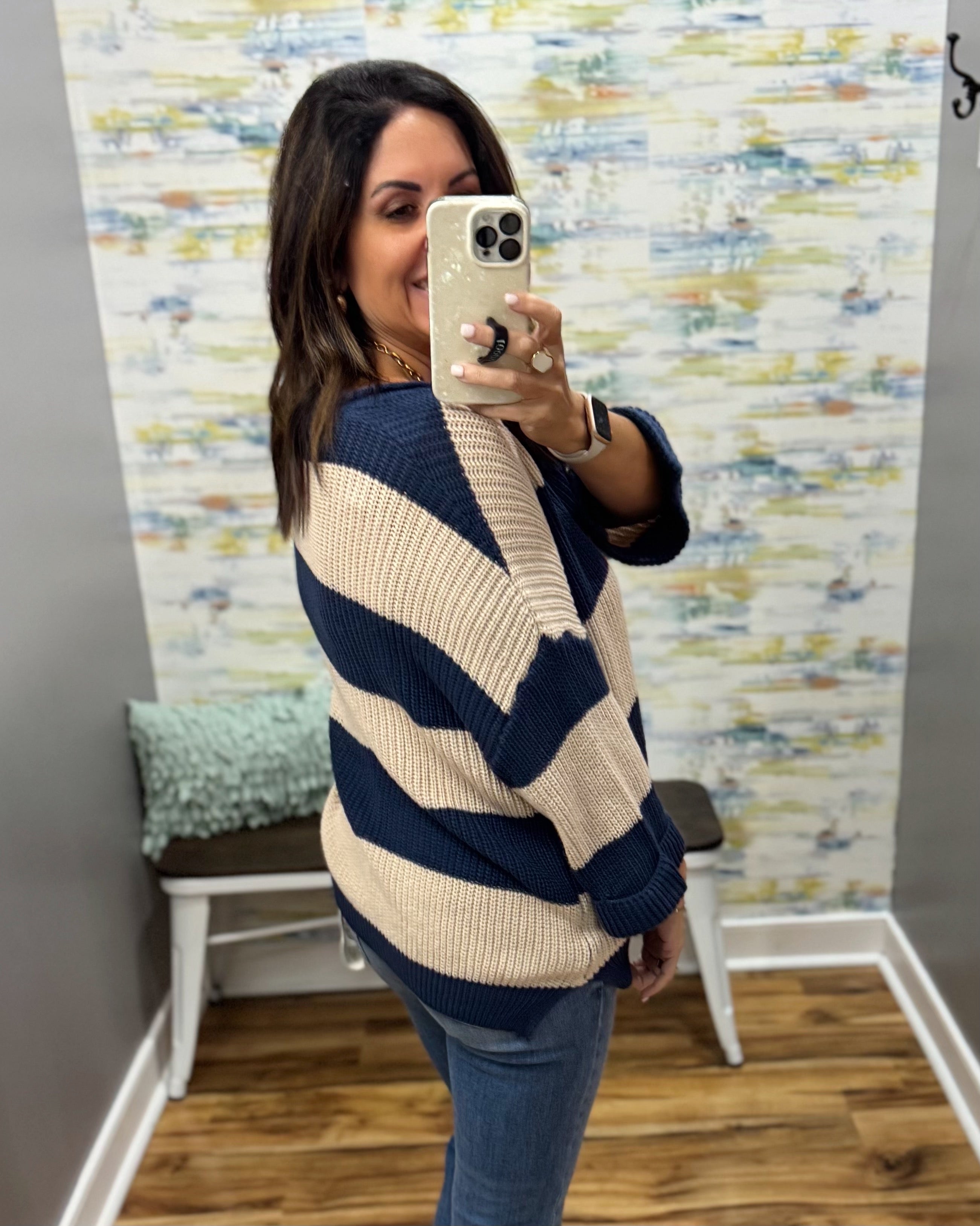A woman takes a mirror selfie wearing a navy blue and cream striped knit sweater, blue jeans, and a gold ring, standing in a room with patterned wallpaper and a wooden floor.