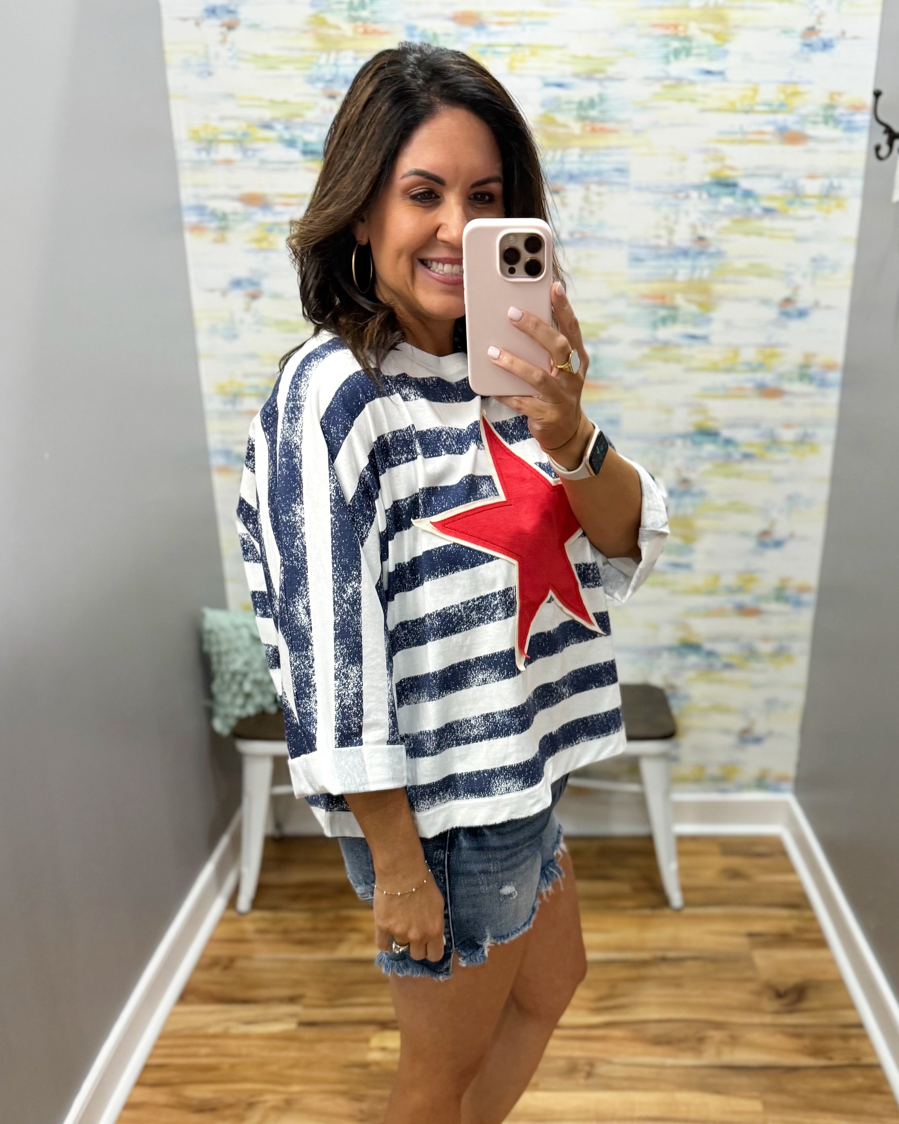 Woman taking a mirror selfie wearing a striped shirt with a red star, standing in a room with a colorful wall.