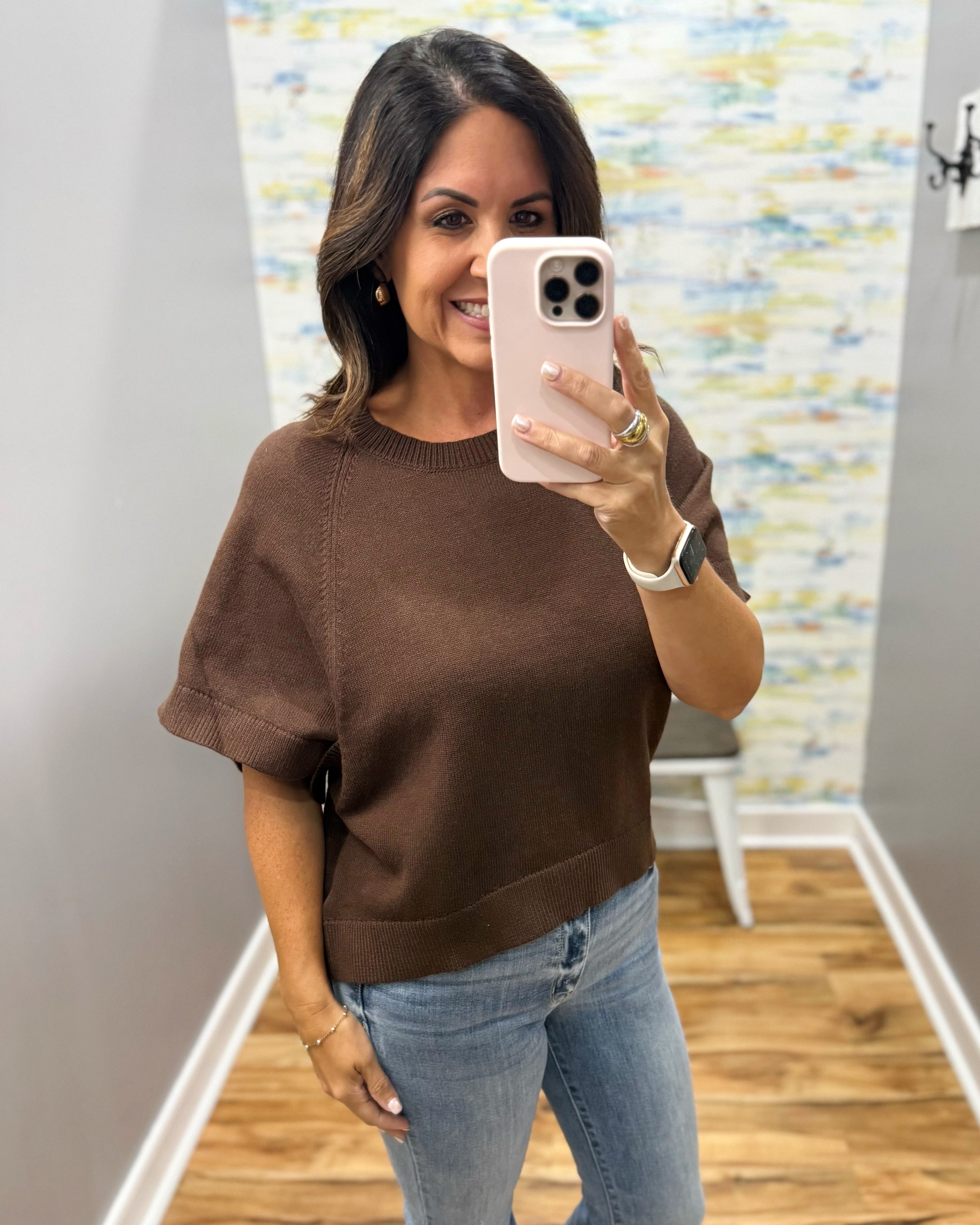 A woman takes a mirror selfie wearing a brown short-sleeve knit sweater and light blue jeans in a home setting with a wood floor and patterned wallpaper.