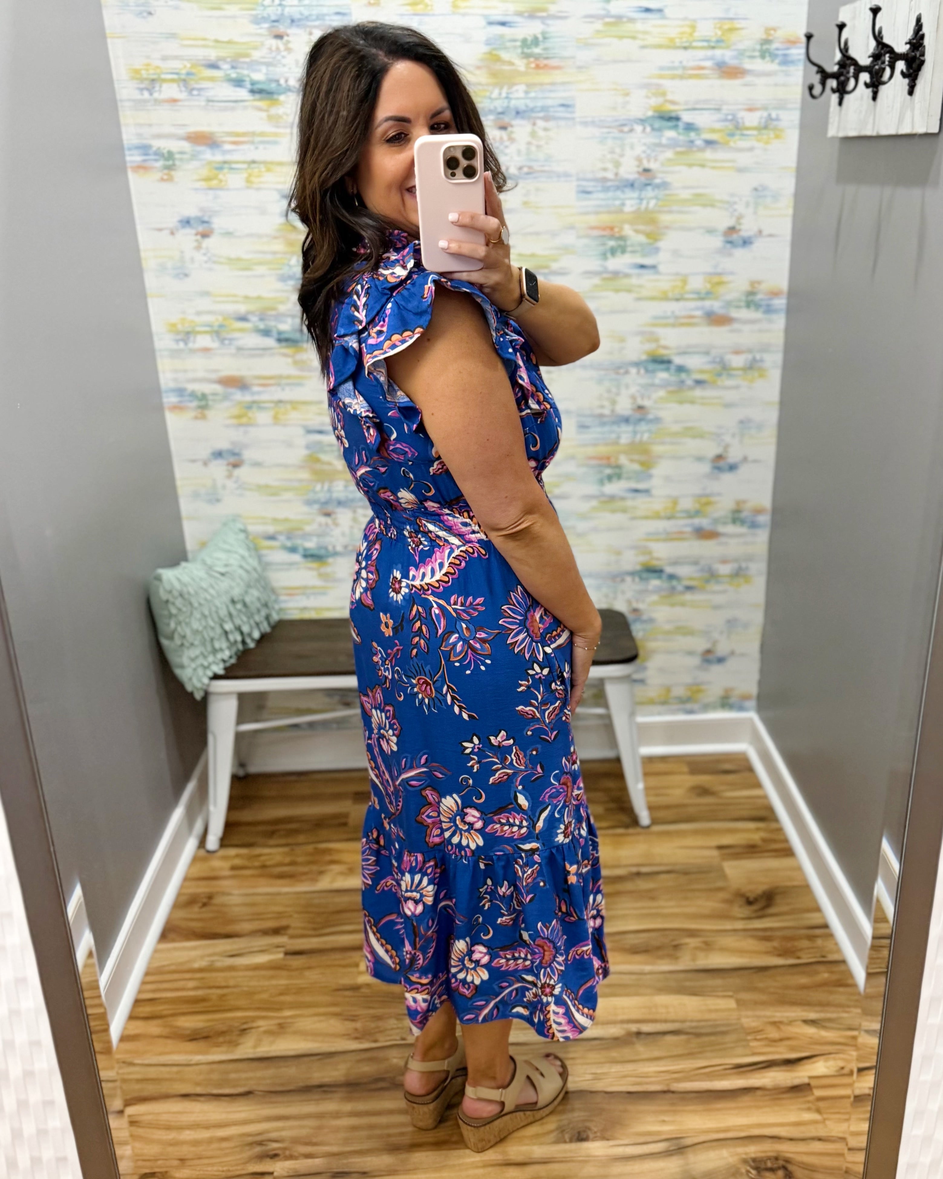 Woman taking a mirror selfie wearing a blue floral dress in a room with patterned wallpaper.