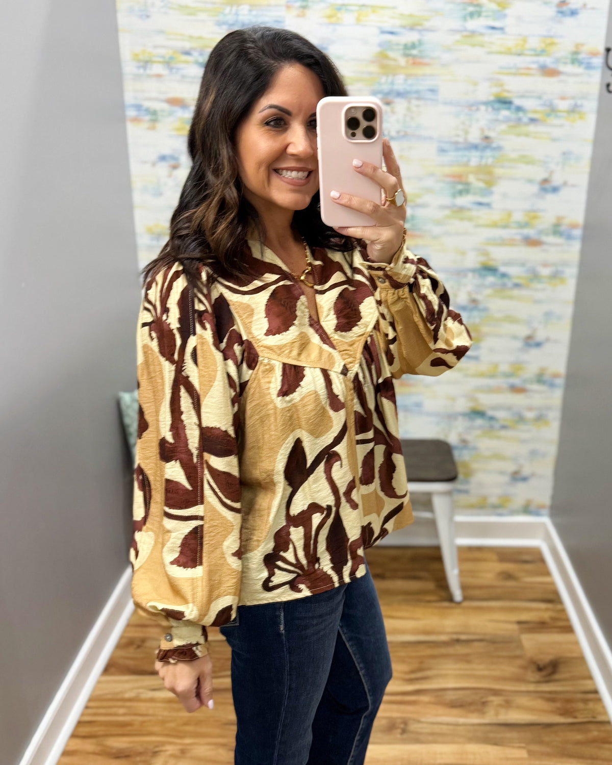 A woman takes a mirror selfie wearing a long-sleeve blouse with a brown and gold abstract botanical pattern, paired with dark blue jeans.
