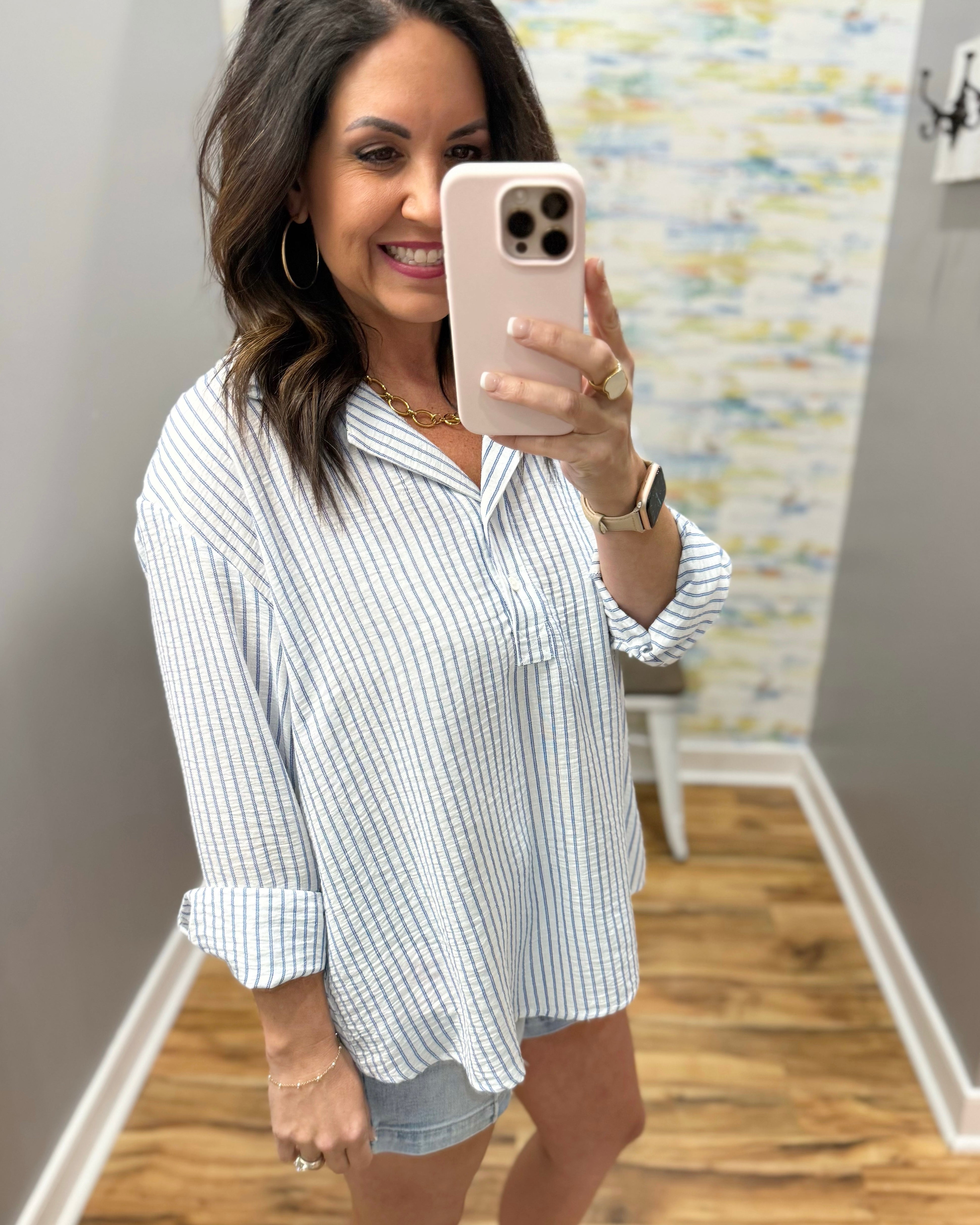 Woman taking a mirror selfie wearing a striped shirt and denim shorts.