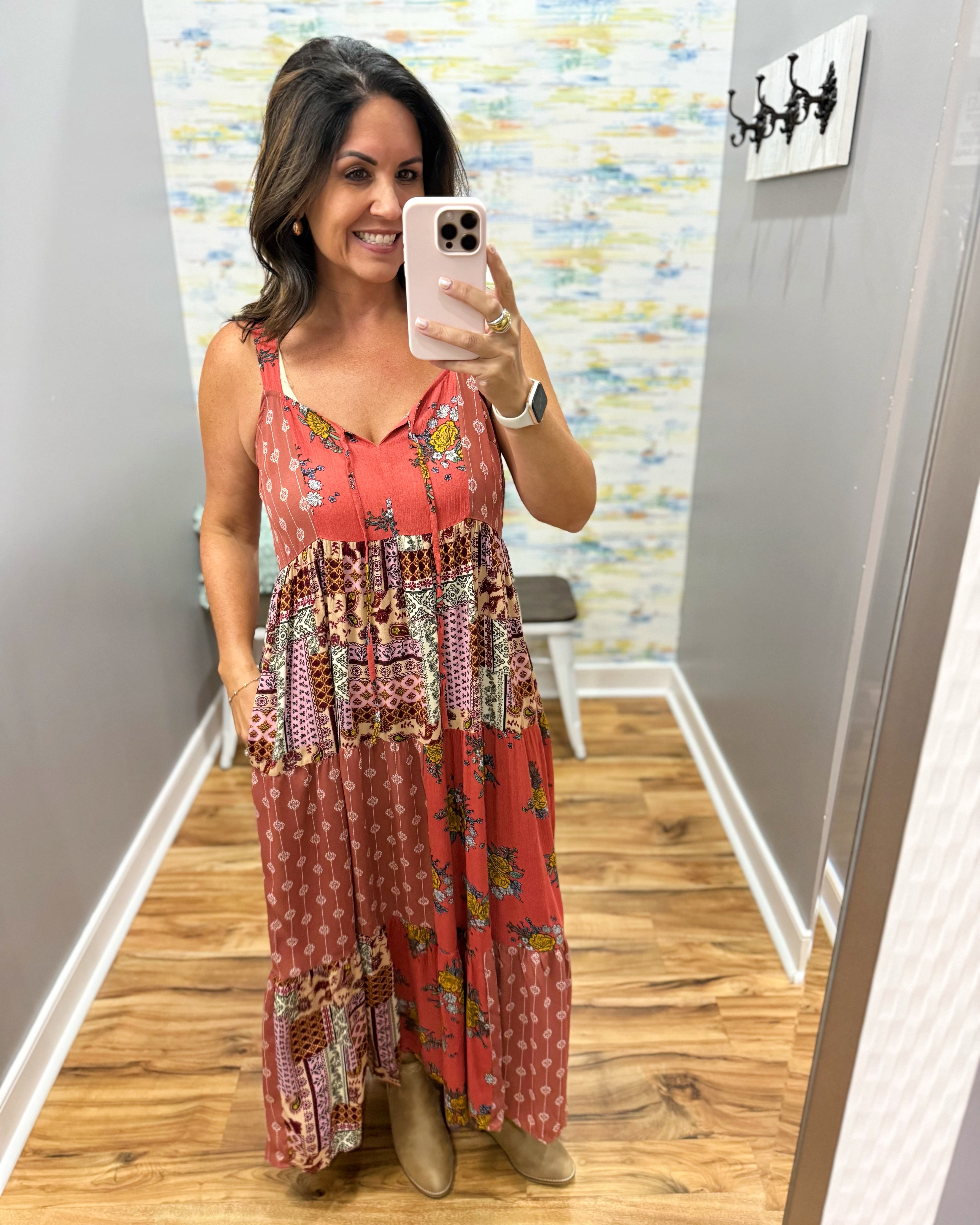 A woman takes a mirror selfie in a fitting room wearing a sleeveless, patchwork-style maxi dress with a rust-red base and multicolor floral and geometric patterns. She is wearing tan boots and holding a light pink smartphone.