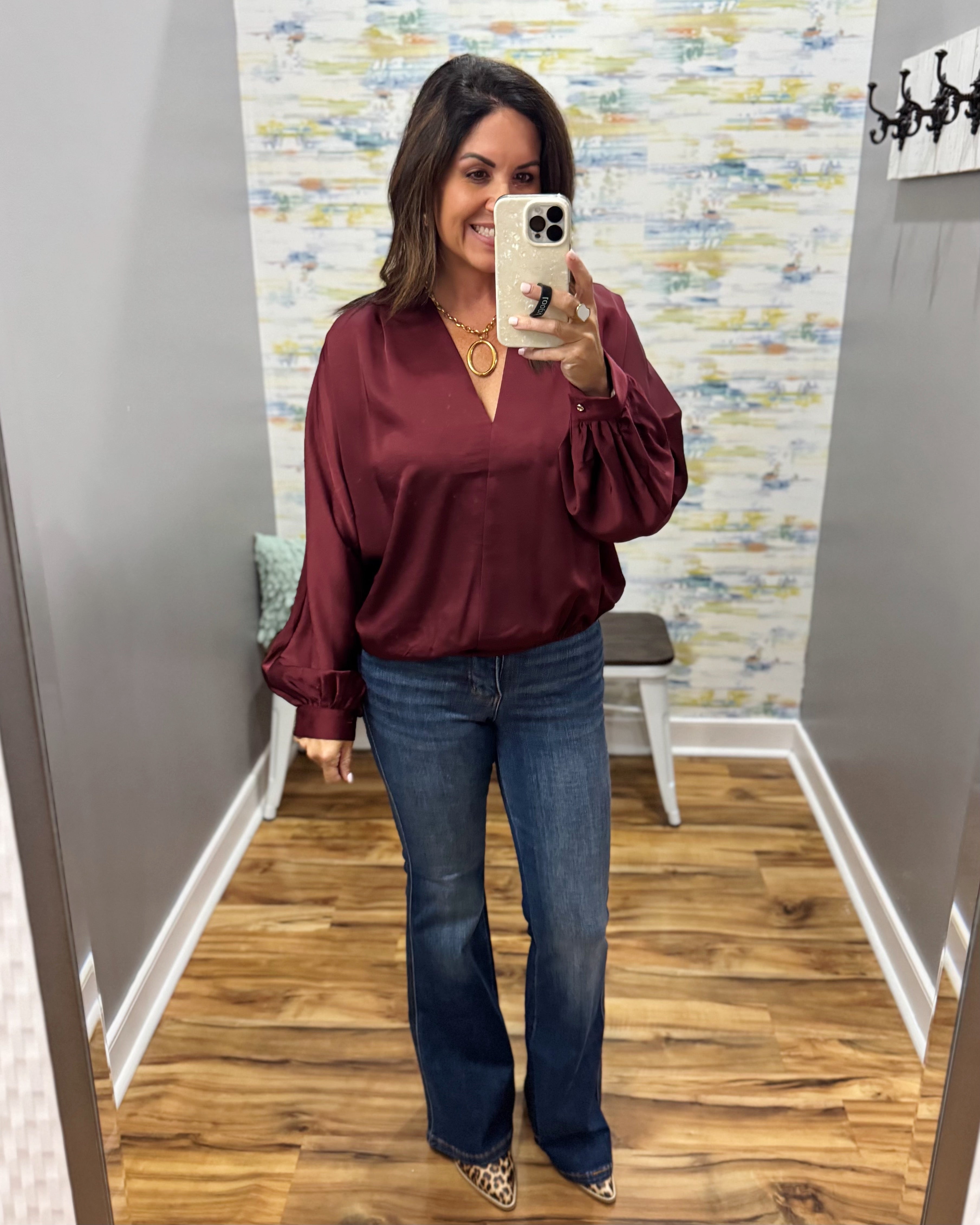 A woman takes a mirror selfie in a fitting room wearing a burgundy satin blouse with balloon sleeves and dark blue flare jeans.