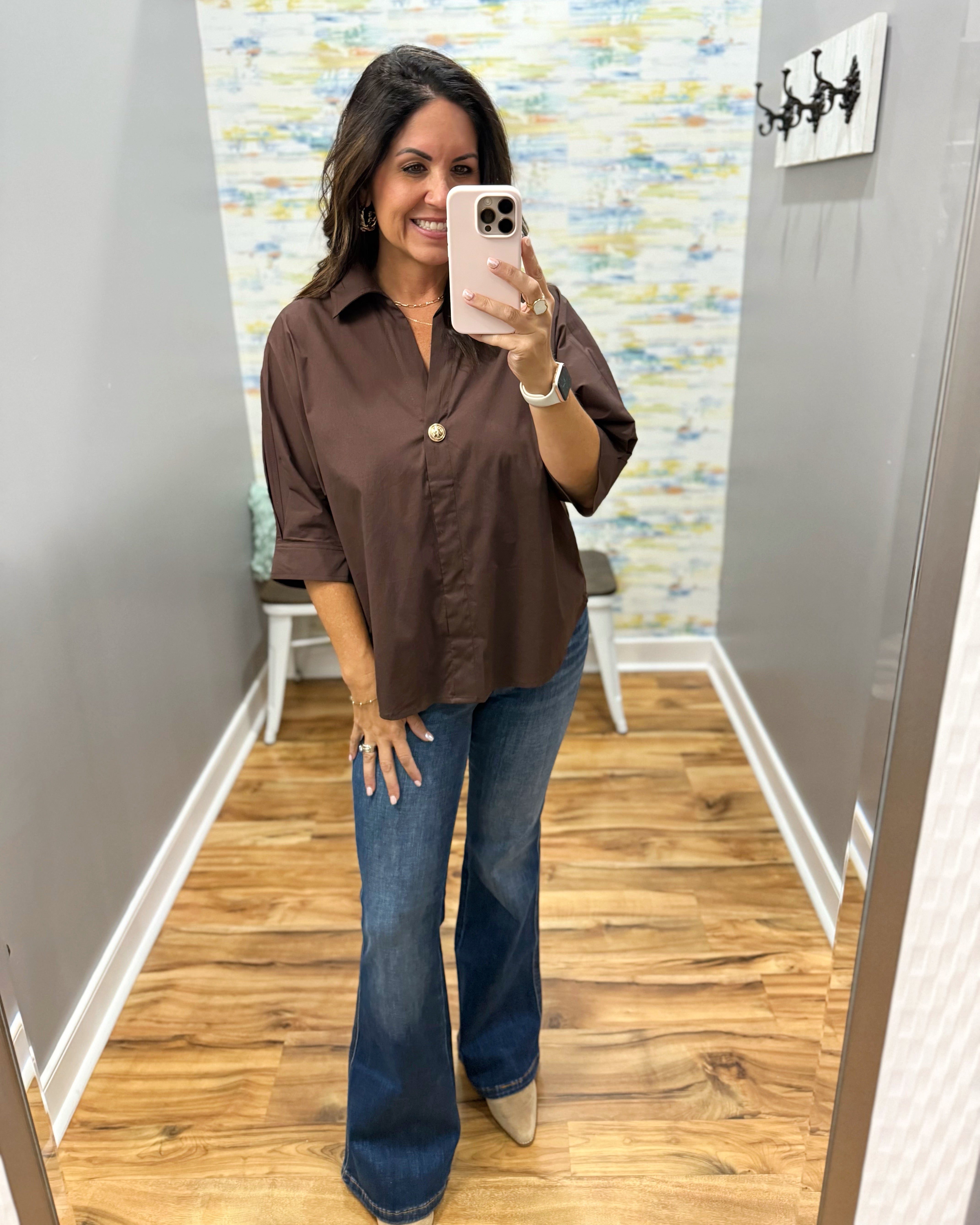 A woman takes a mirror selfie in a fitting room wearing a brown button-down shirt with rolled sleeves and blue flare jeans.