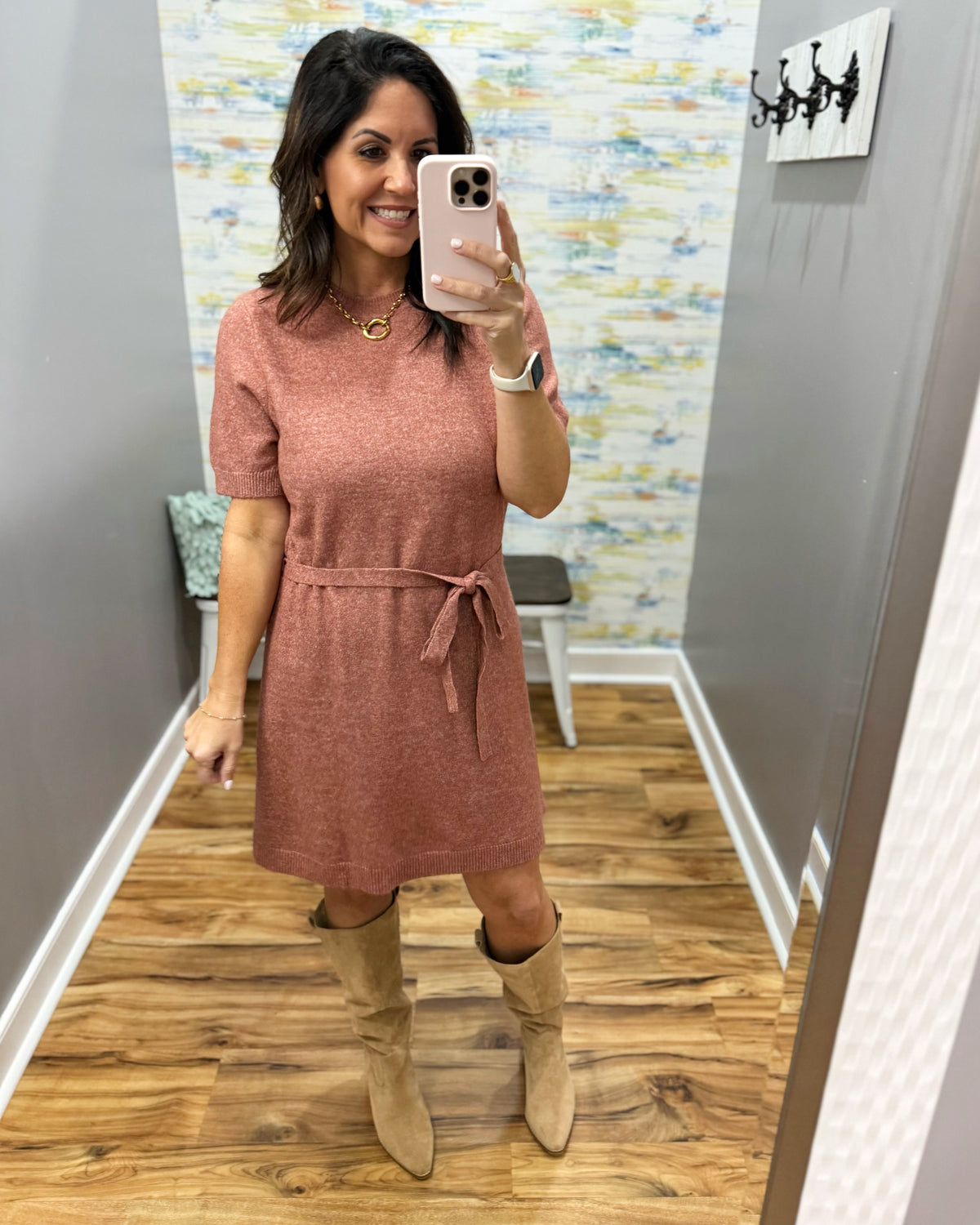 A woman takes a mirror selfie wearing a short-sleeve dusty rose knit dress with a tie waist and tan knee-high boots in a fitting room.