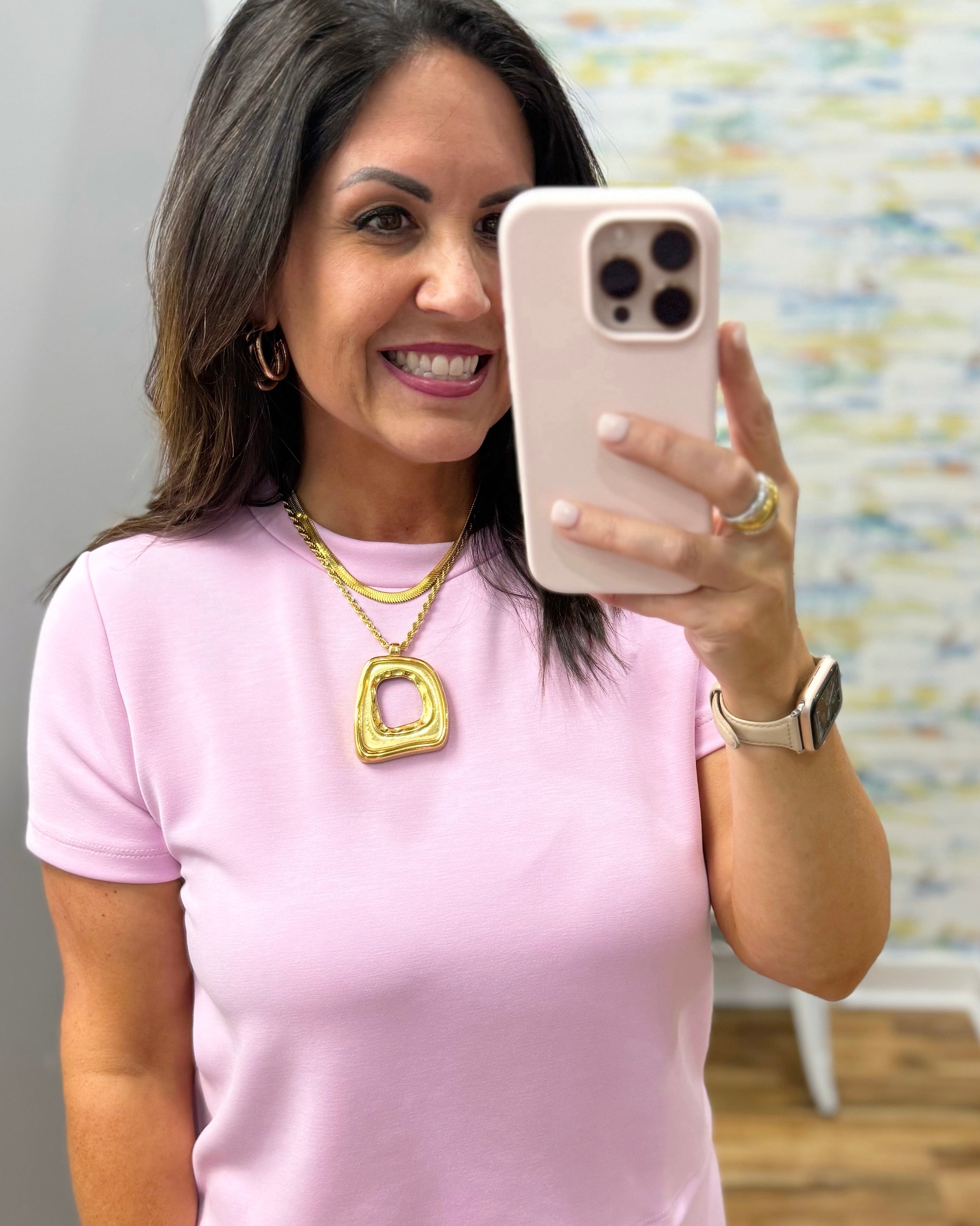 Woman in a pink shirt taking a selfie with a phone, wearing gold jewelry.