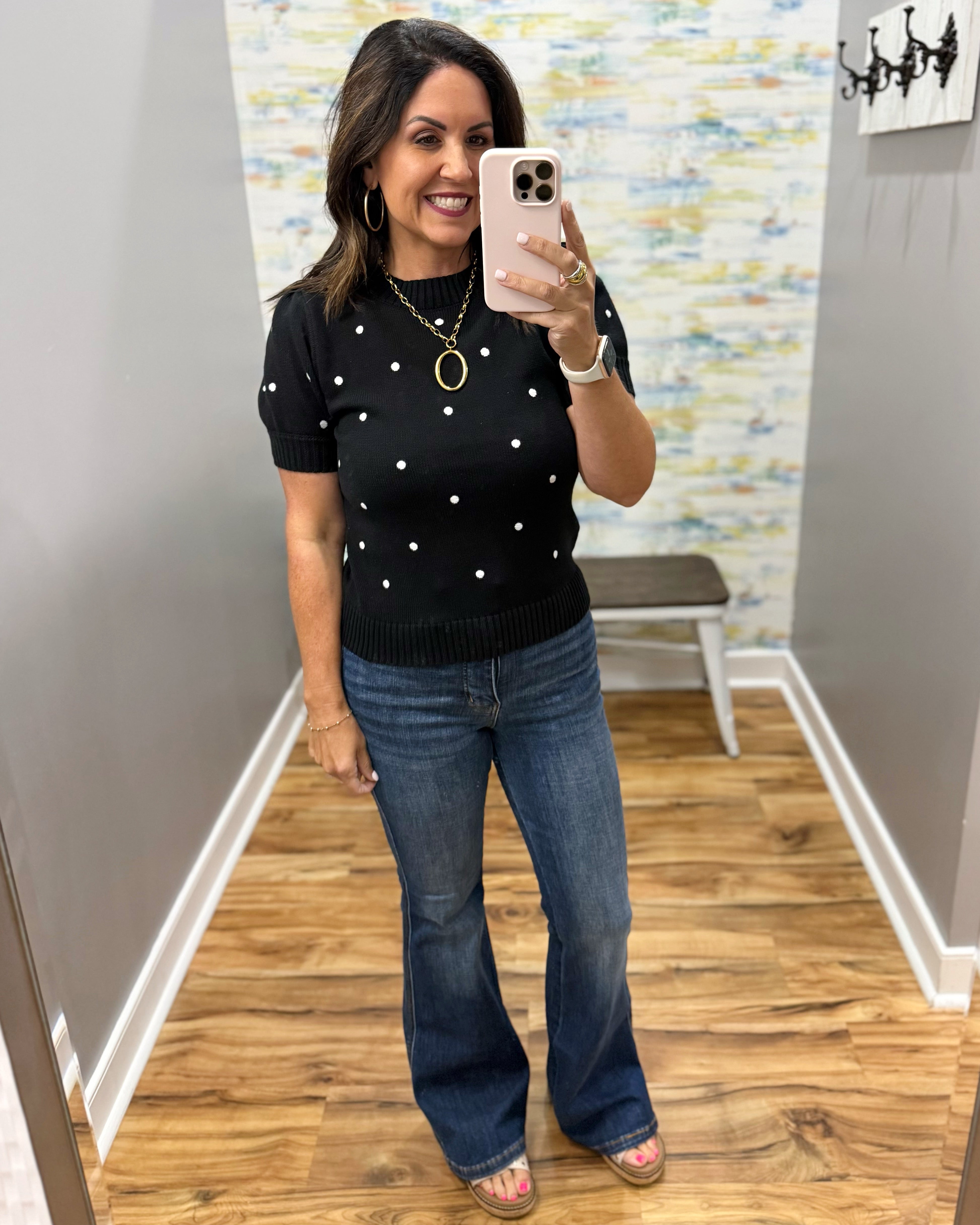 A woman takes a mirror selfie in a fitting room wearing a black short-sleeve knit sweater with white polka dots, blue flare jeans, and flat sandals.