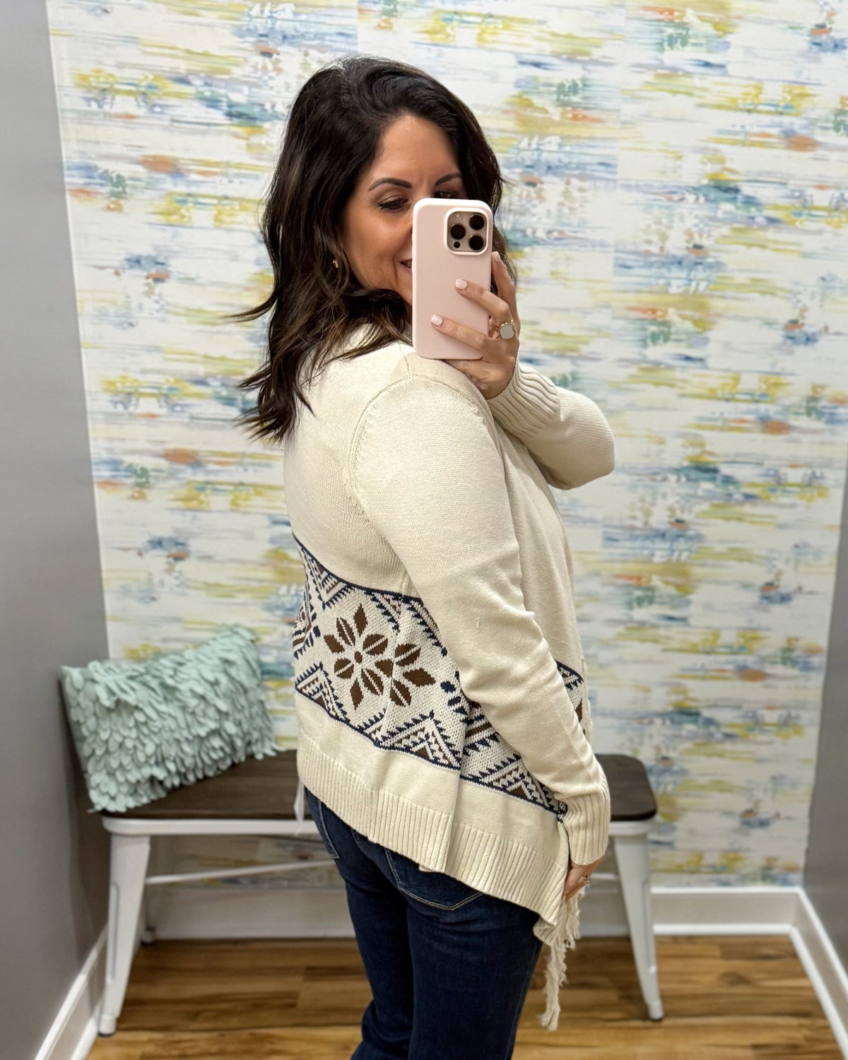 A woman takes a mirror selfie in a fitting room wearing a cream knit cardigan with a brown and black geometric pattern across the back and sleeves, paired with dark blue jeans.