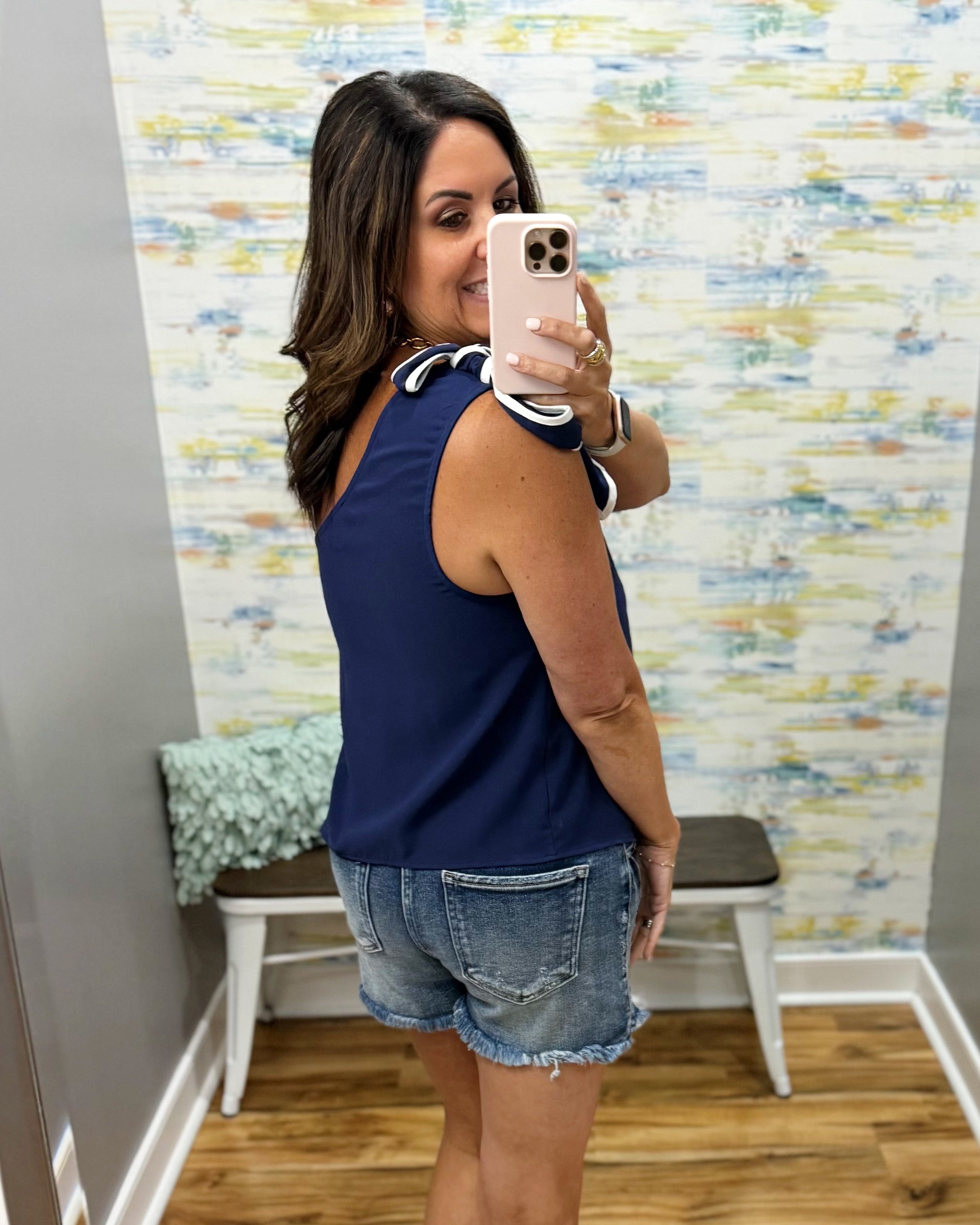 Woman taking a mirror selfie wearing a navy tank top and denim shorts in a room with patterned wallpaper.