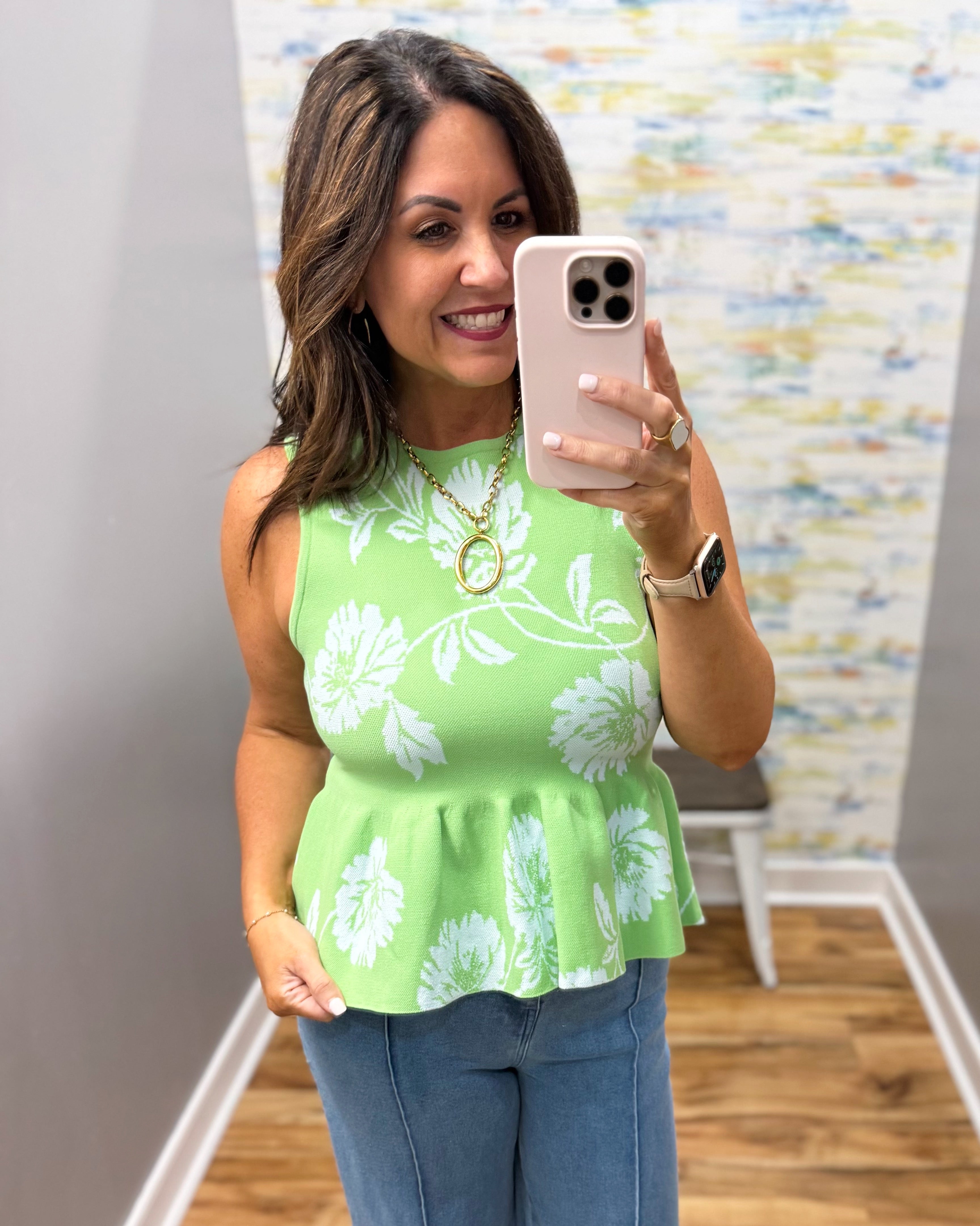 Woman taking a mirror selfie wearing a green floral top and blue jeans.