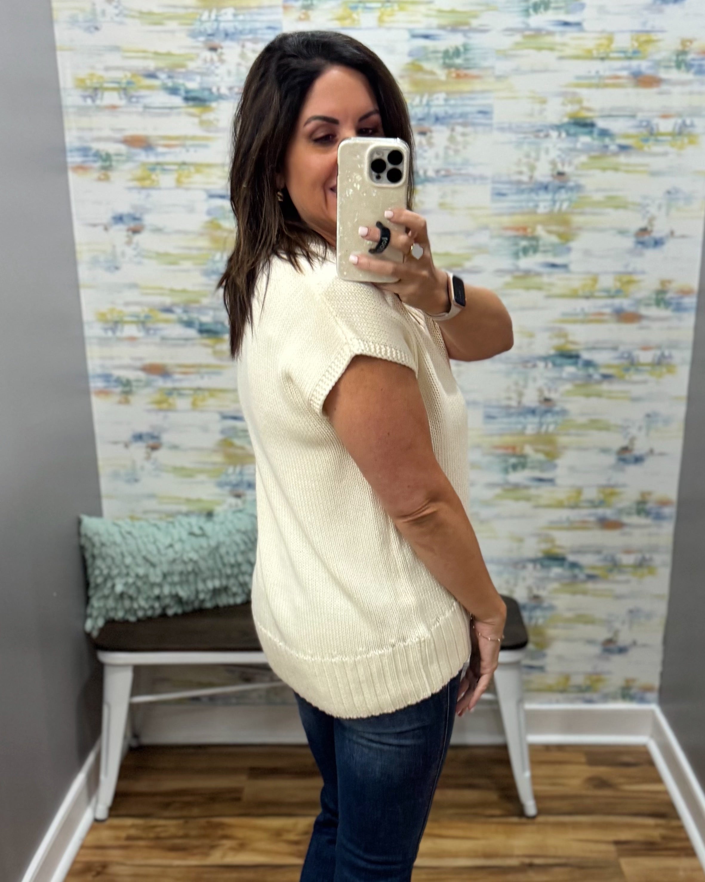 A woman takes a mirror selfie in a fitting room wearing a cream-colored short-sleeve knit sweater vest and dark blue jeans. The background features a patterned wallpaper and a bench with a green pillow.