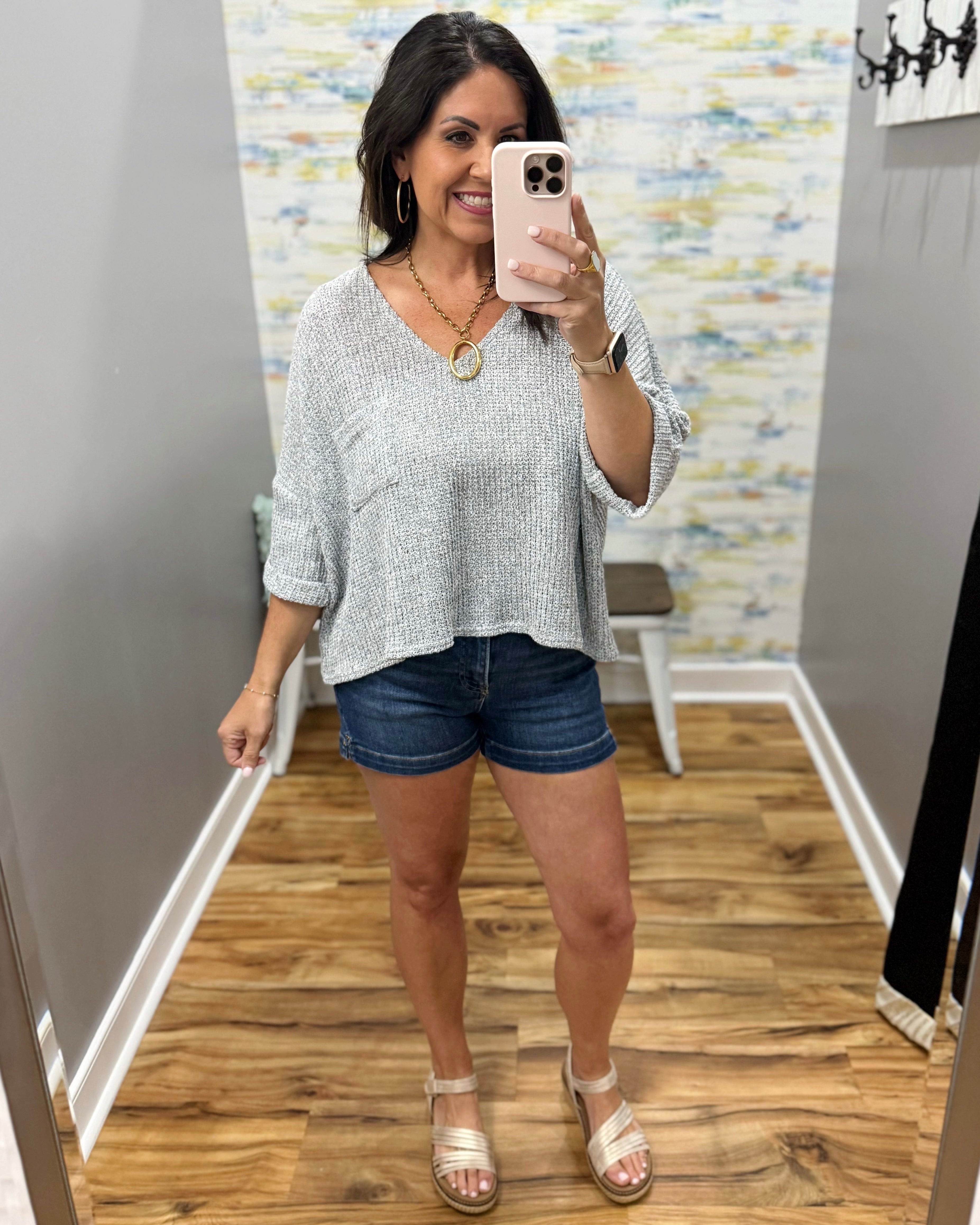 Woman taking a mirror selfie wearing a light gray sweater and denim shorts in a room with a patterned wall.
