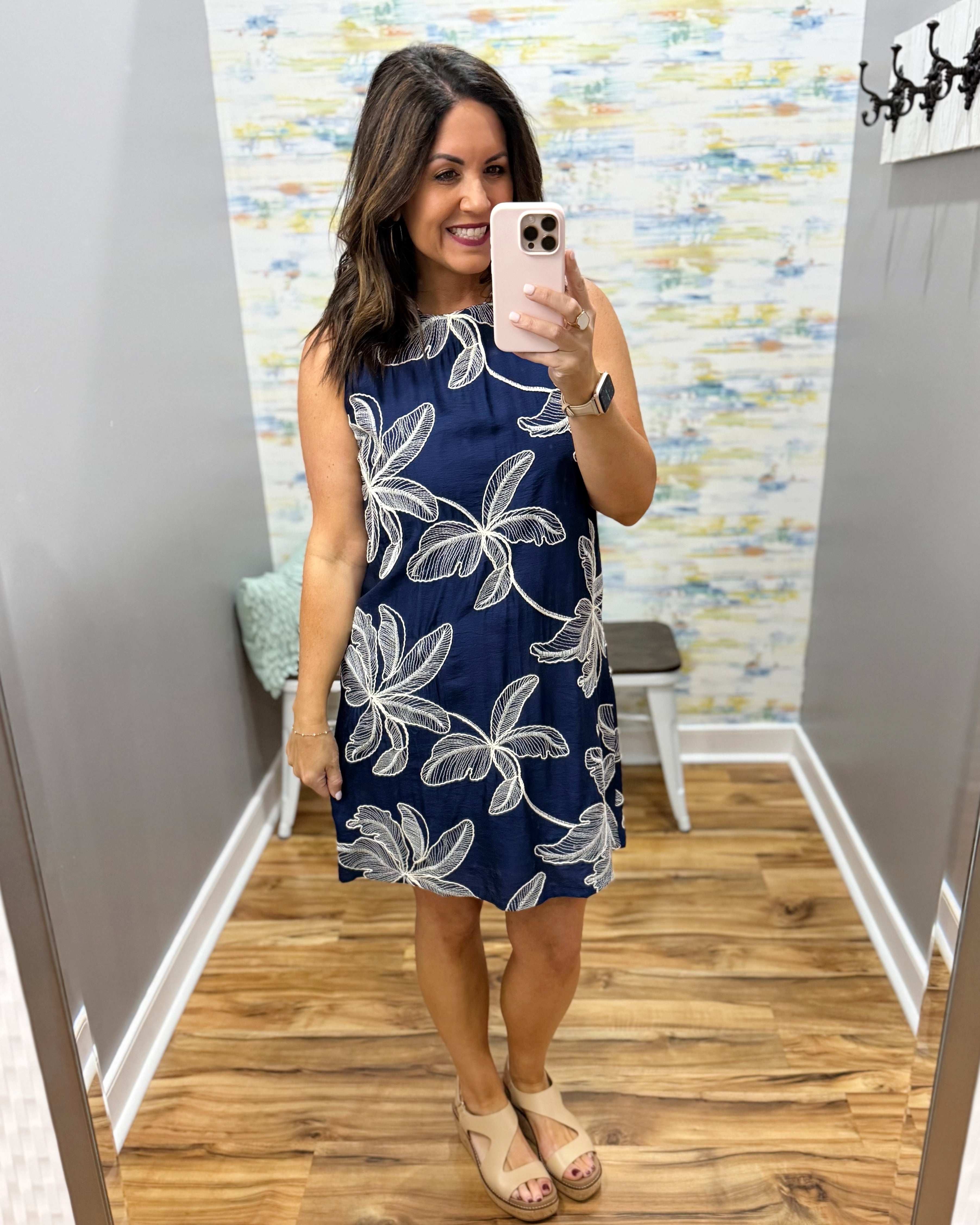 Woman taking a mirror selfie wearing a navy dress with white floral patterns in a room with a patterned wall.