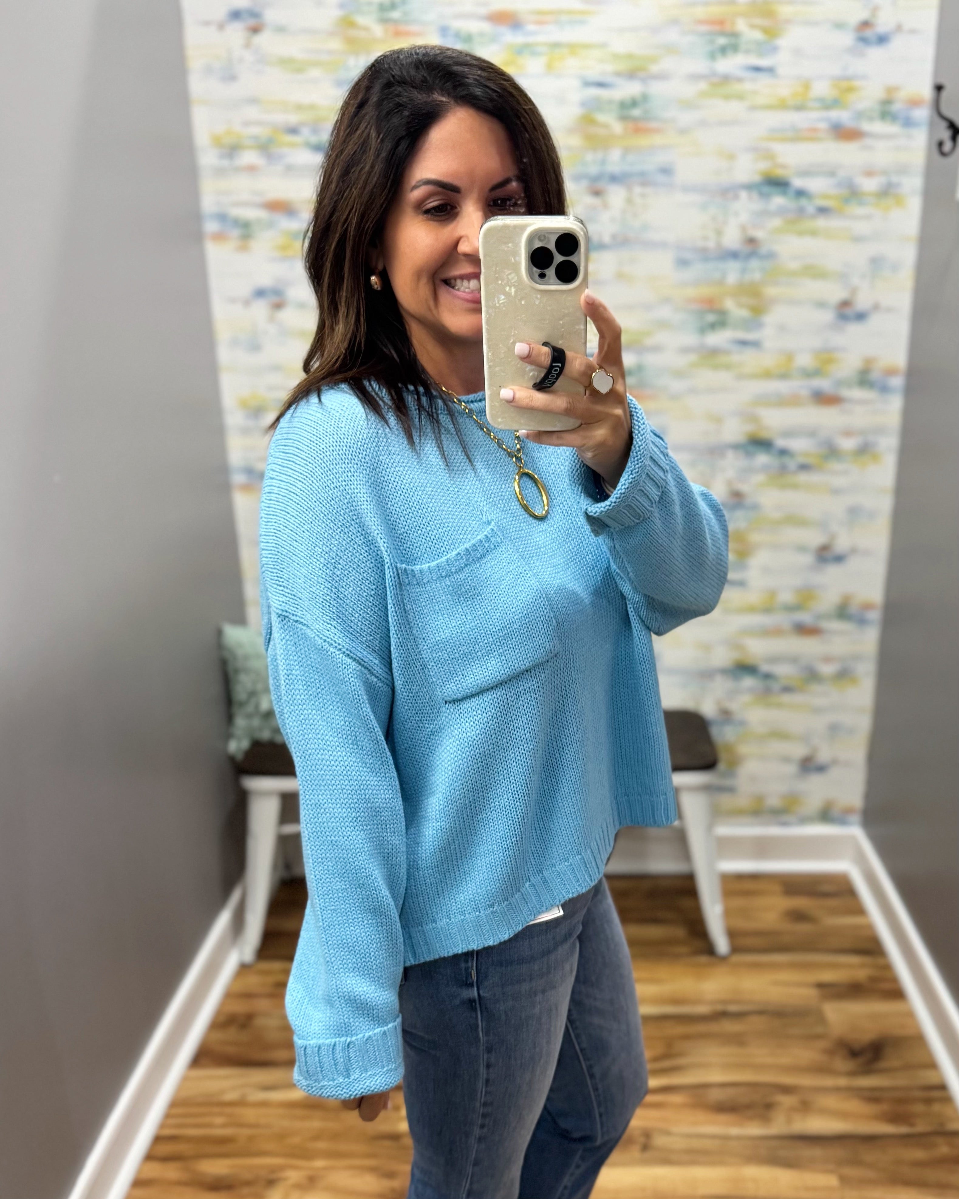A woman takes a mirror selfie wearing a light blue knit sweater with a chest pocket and dark gray jeans in a fitting room with a patterned wallpaper background.