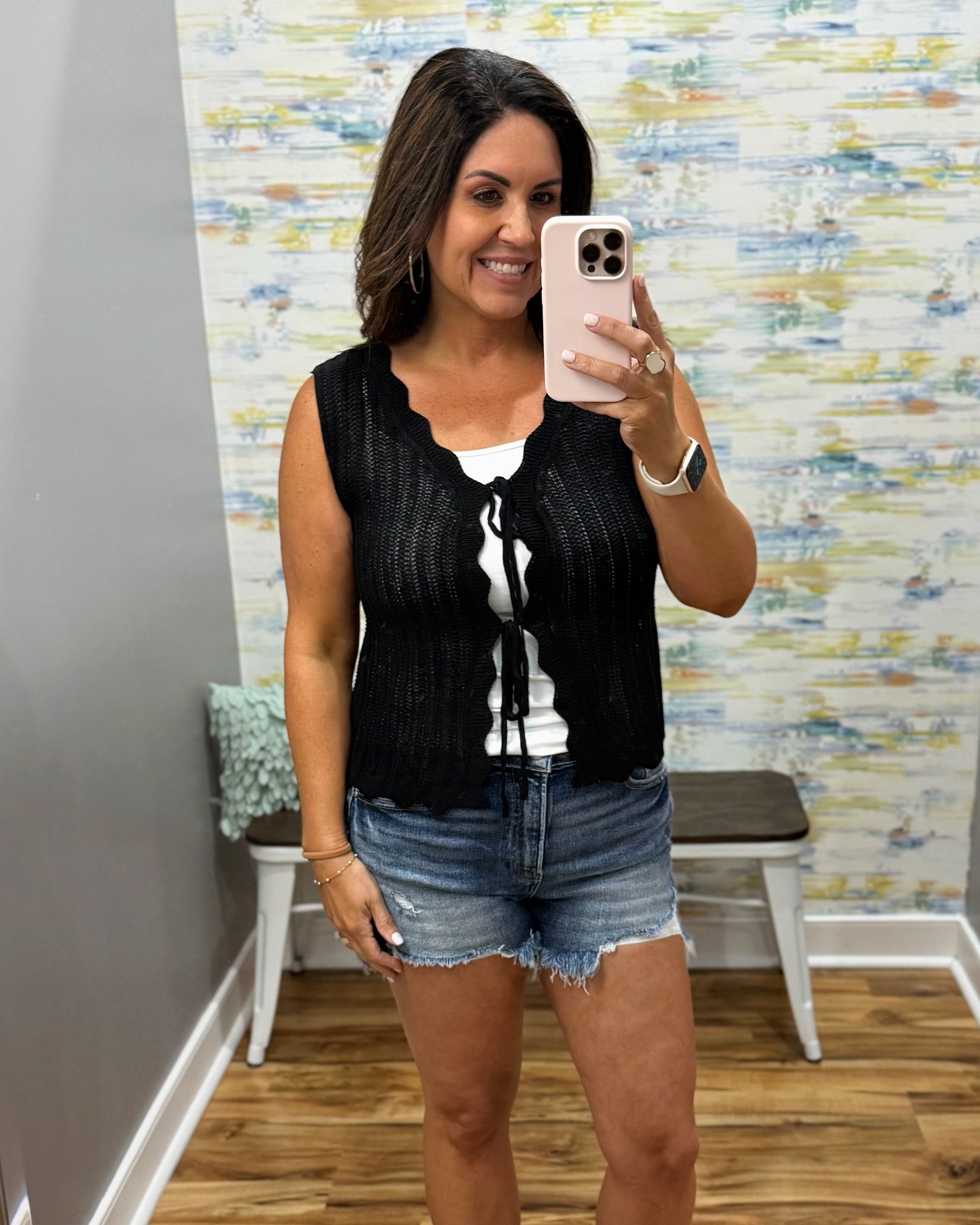 Woman taking a mirror selfie wearing a black sleeveless top and denim shorts in a room with a textured wall.