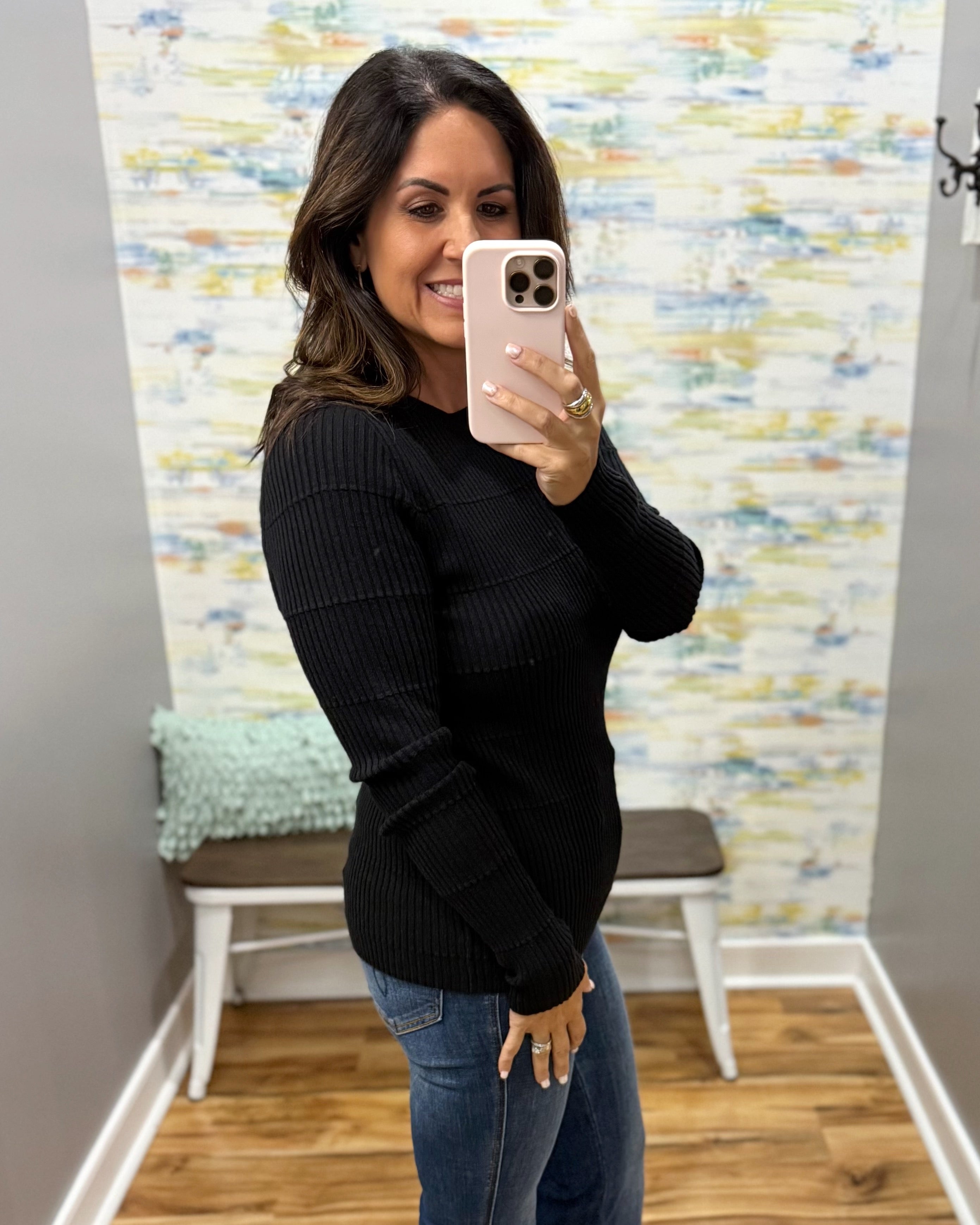 A woman takes a mirror selfie wearing a black ribbed long-sleeve knit sweater and blue jeans in an indoor room with a patterned wallpaper background.