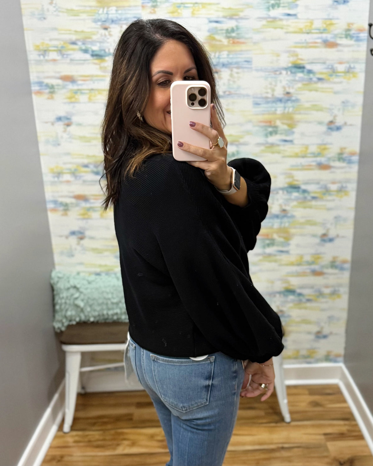 A woman takes a mirror selfie wearing a black long-sleeve cropped sweater and light blue jeans, standing in front of a patterned wallpaper background.