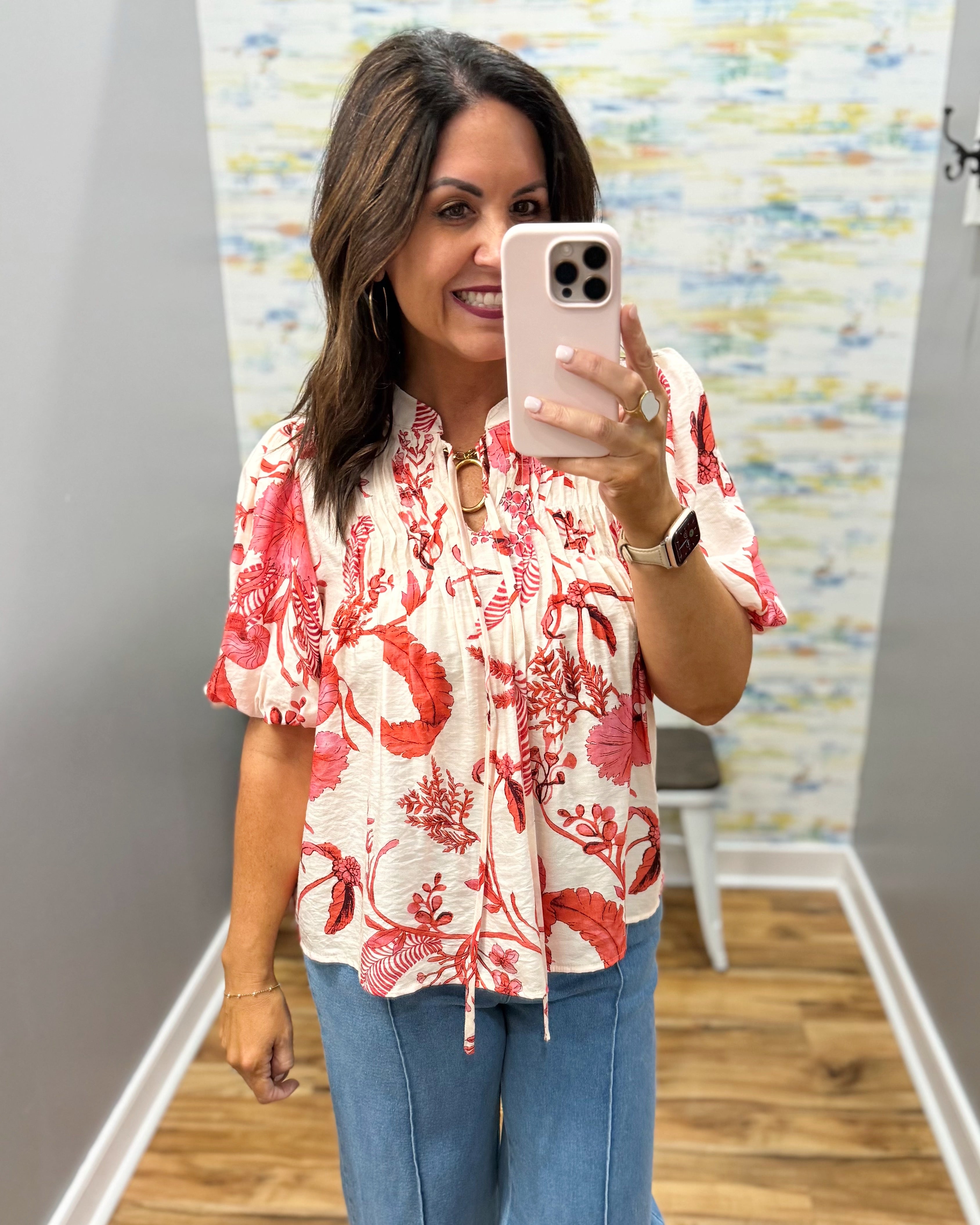 Woman taking a mirror selfie wearing a red floral blouse and blue jeans in a room with a colorful wall.
