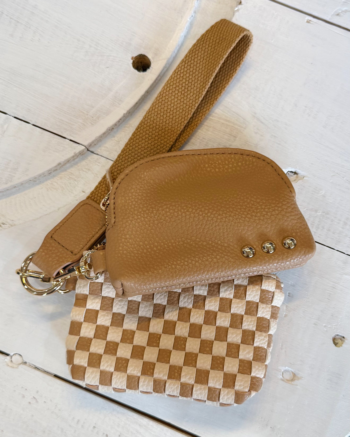 A tan pebbled leather crossbody bag with a wide strap, laid on a white wooden surface next to a woven checkered pouch.