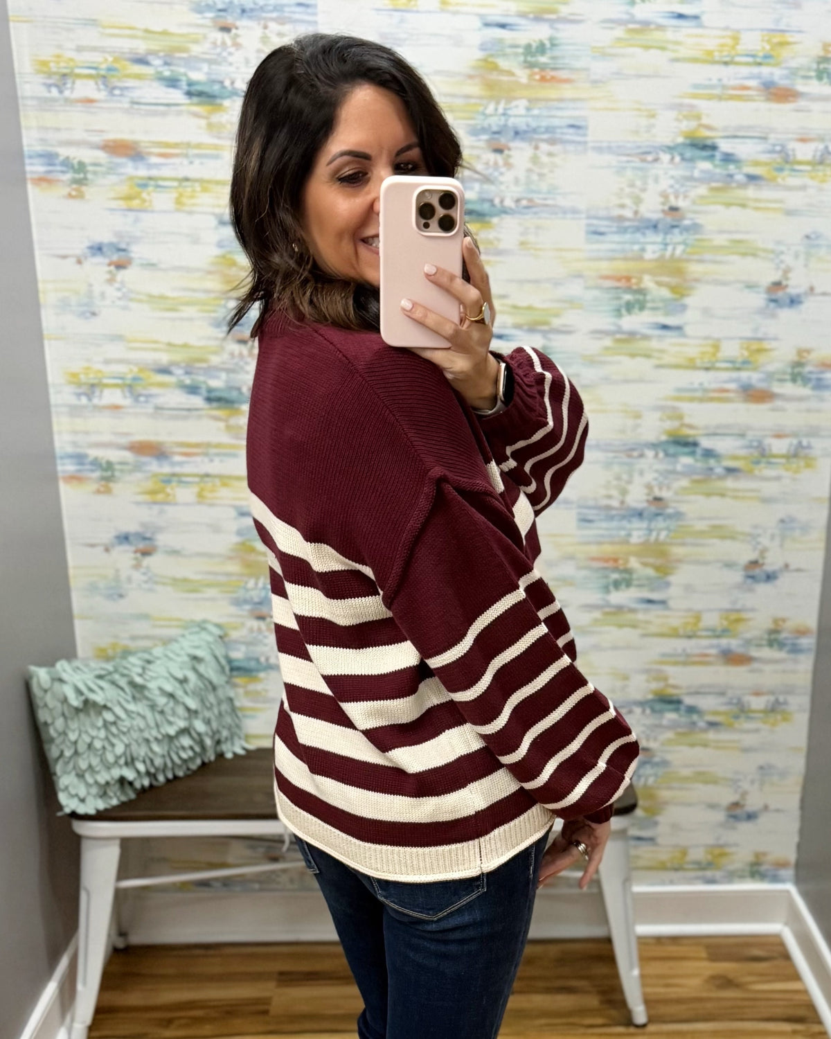 A woman takes a mirror selfie wearing a color-block knit sweater with a burgundy and cream horizontal stripe pattern, paired with dark blue jeans. She is standing in front of a patterned wallpaper background.