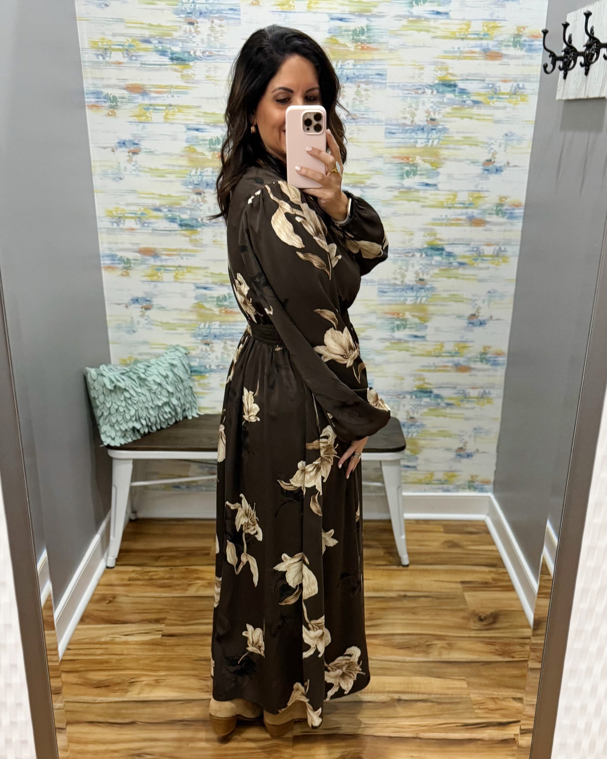 A woman takes a mirror selfie in a boutique fitting room wearing a long-sleeve brown maxi dress with a beige floral pattern. The room features a colorful abstract wallpaper, a wooden floor, and a small bench with a mint green pillow.