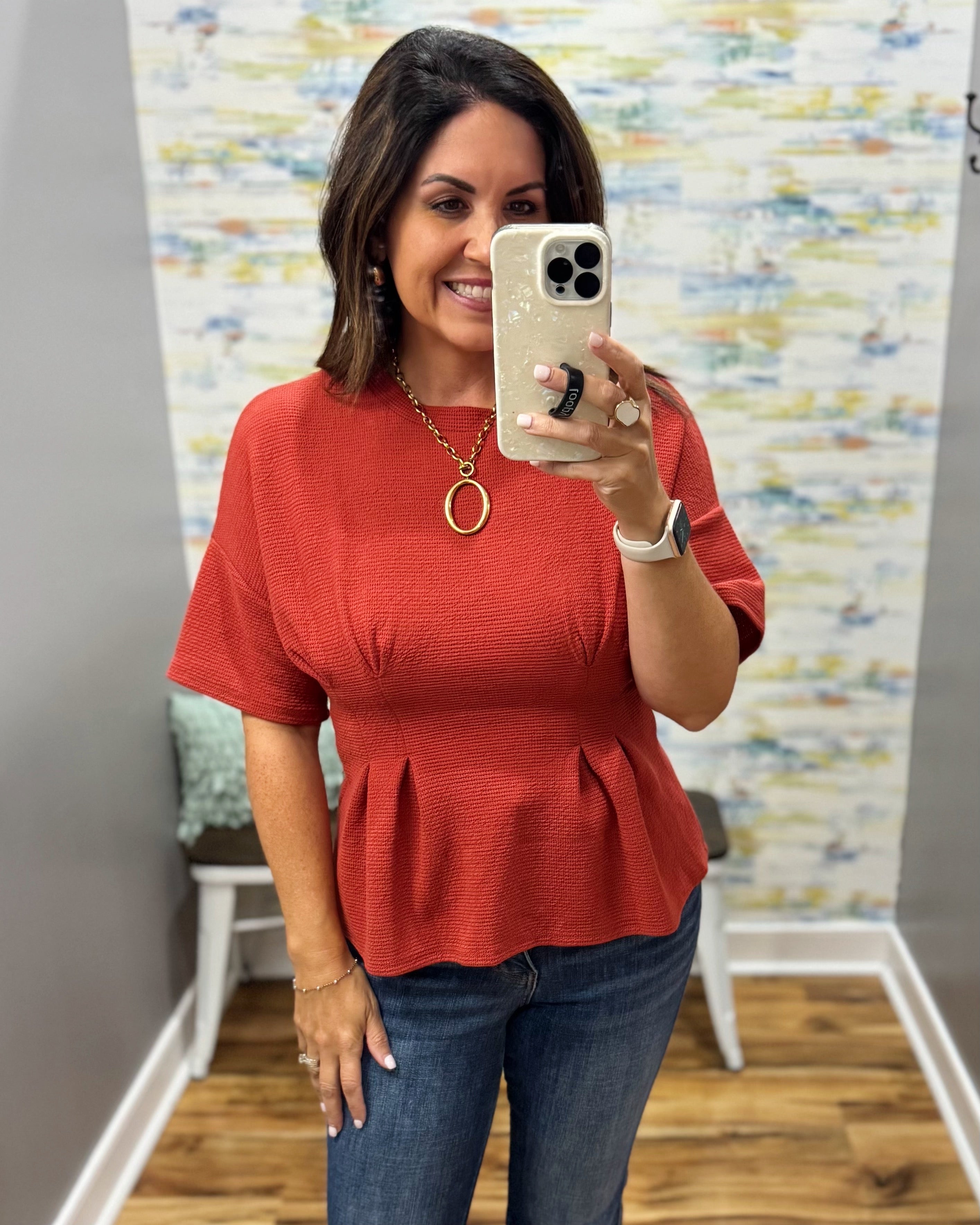 A woman takes a mirror selfie in a fitting room wearing a textured short-sleeve peplum top in a rust red color, paired with blue jeans.