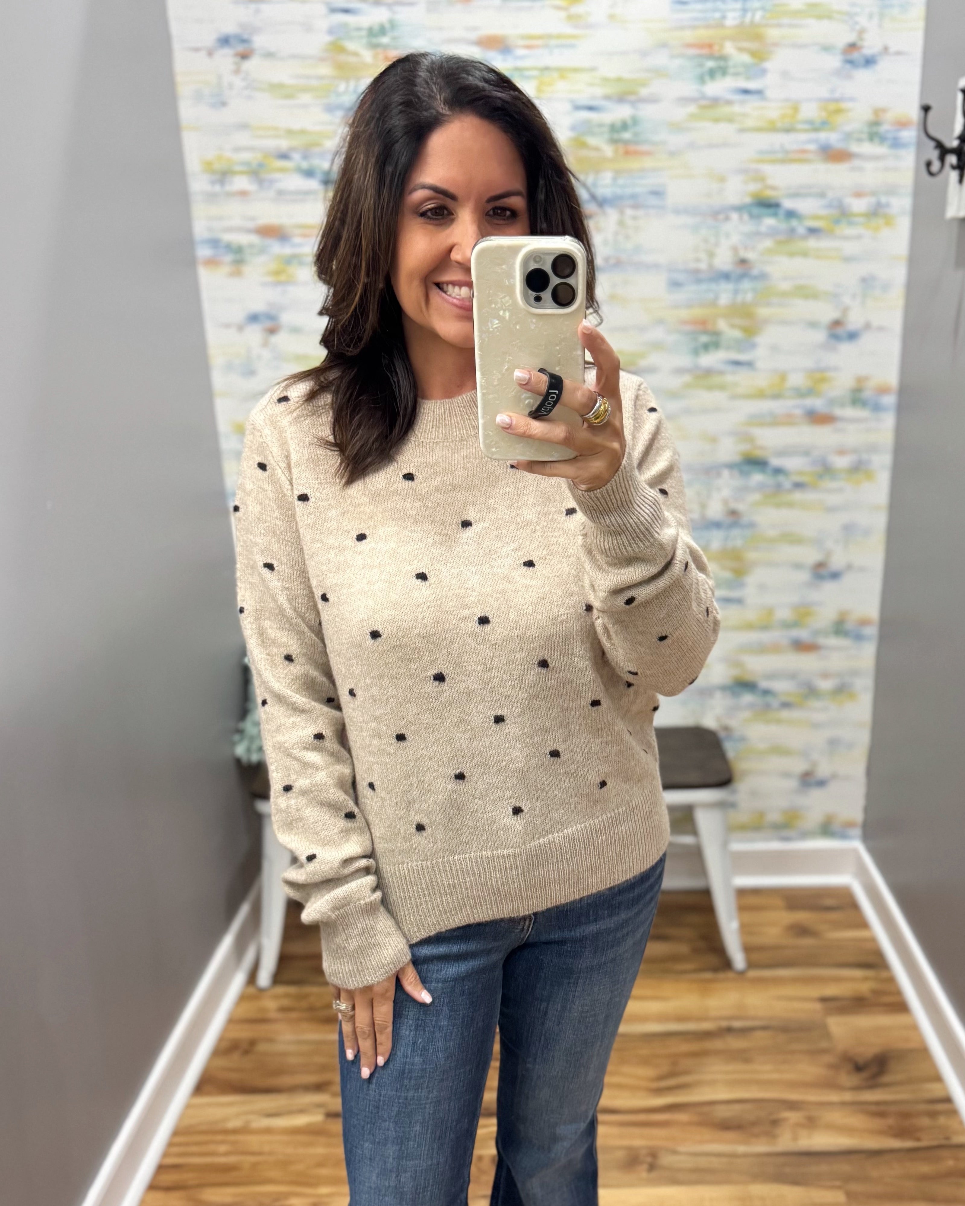 A woman takes a mirror selfie wearing a beige knit sweater with small black embroidered dots and blue jeans in a home setting with a patterned wallpaper background.