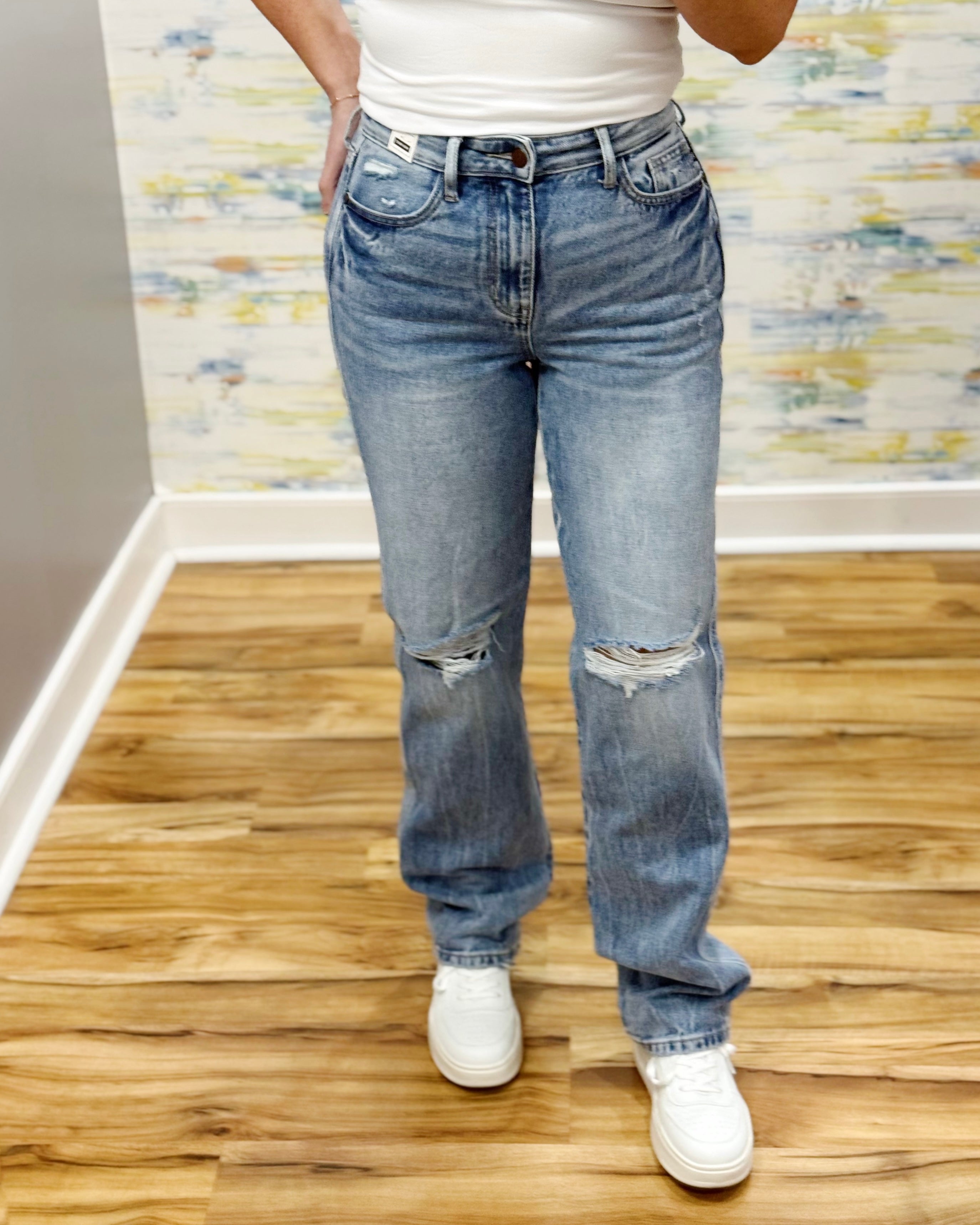 Person wearing blue jeans and white sneakers on a wooden floor with a patterned wall in the background