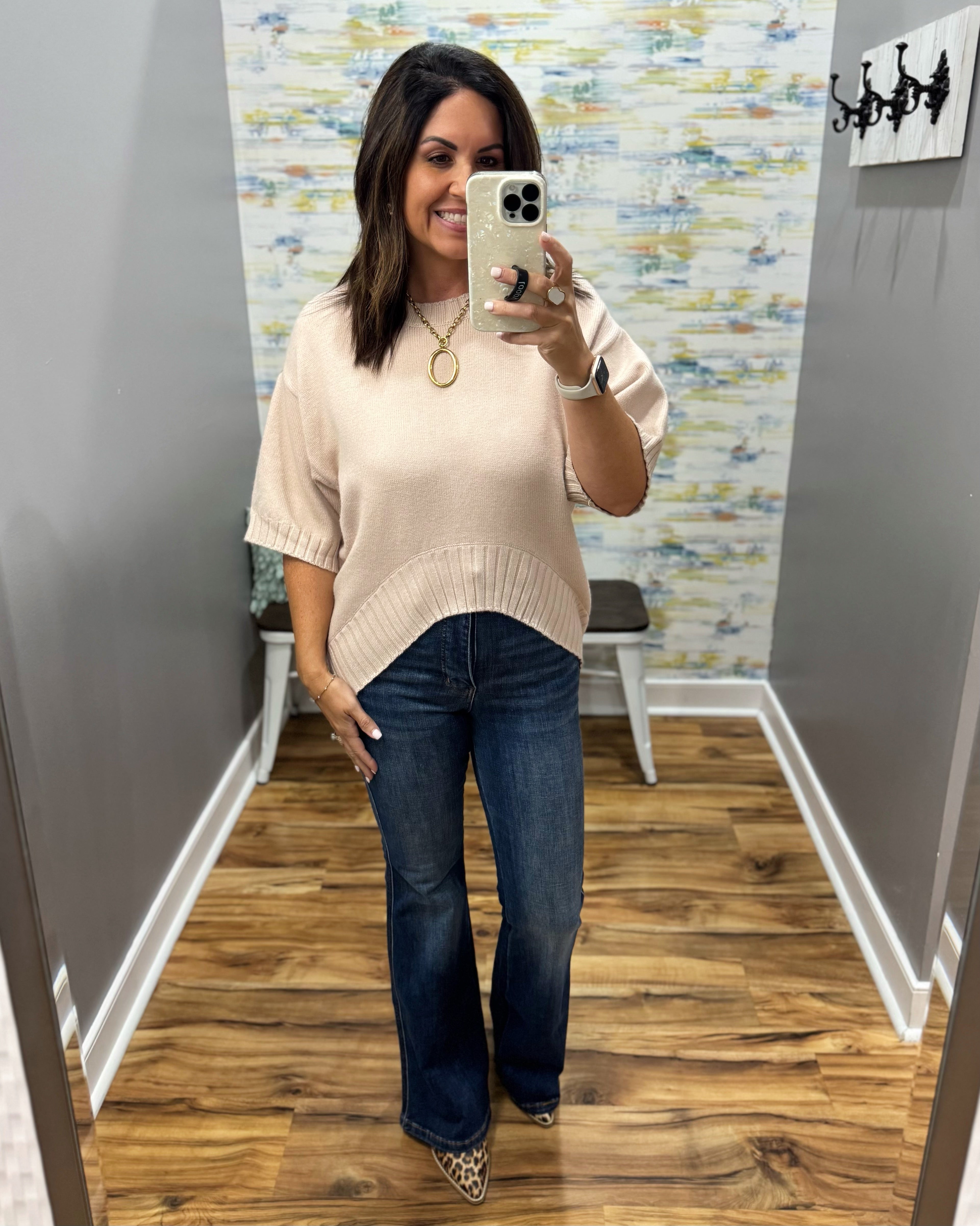 A woman takes a mirror selfie in a fitting room wearing a light beige short-sleeve knit top with a curved hem, dark blue flare jeans, and leopard print heels.
