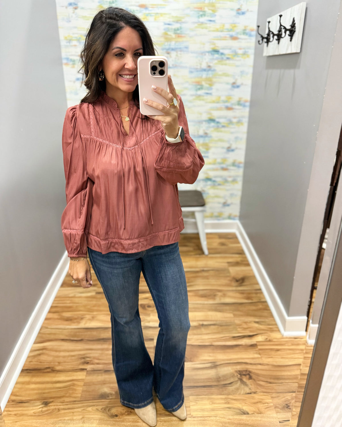 A woman takes a mirror selfie in a boutique fitting room, wearing a dusty rose satin long-sleeve blouse with puff sleeves and blue flare jeans.