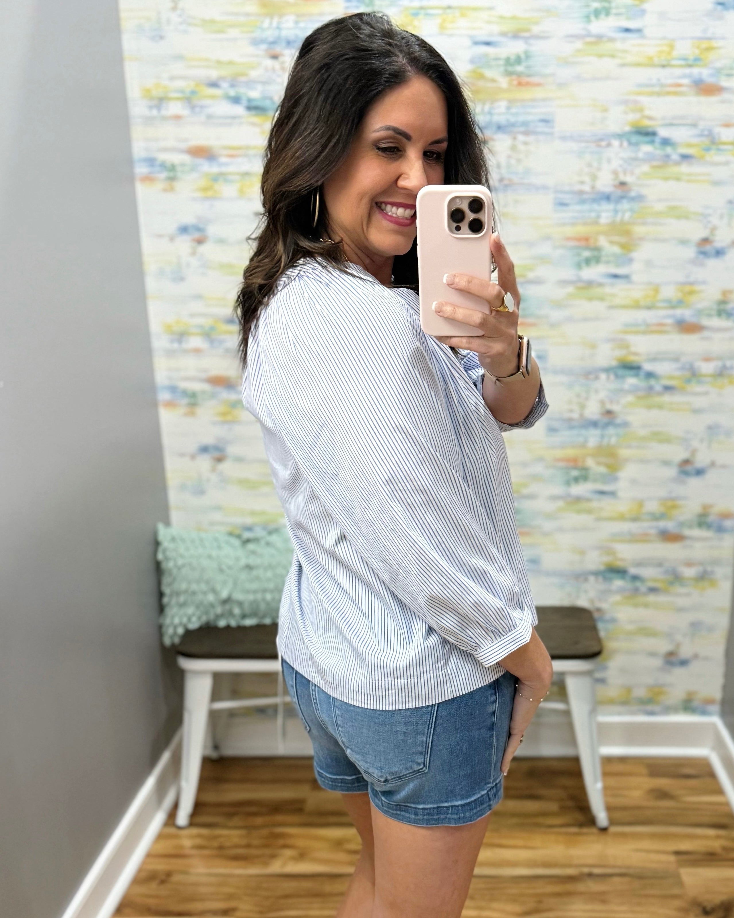 Woman taking a mirror selfie wearing a white striped shirt and denim shorts in a room with colorful wall art.