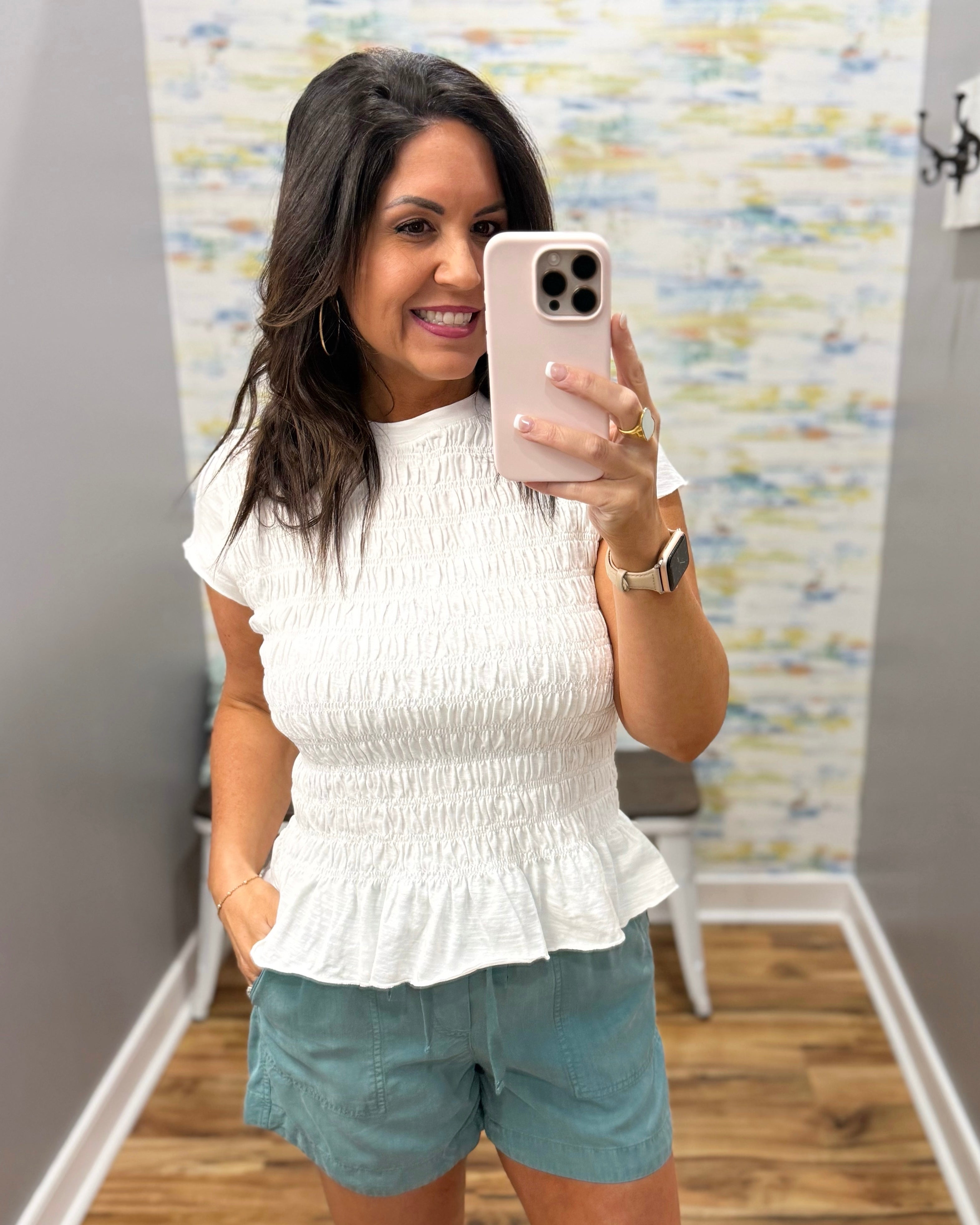 Woman taking a mirror selfie wearing a white top and light blue shorts in a room with colorful wall art.