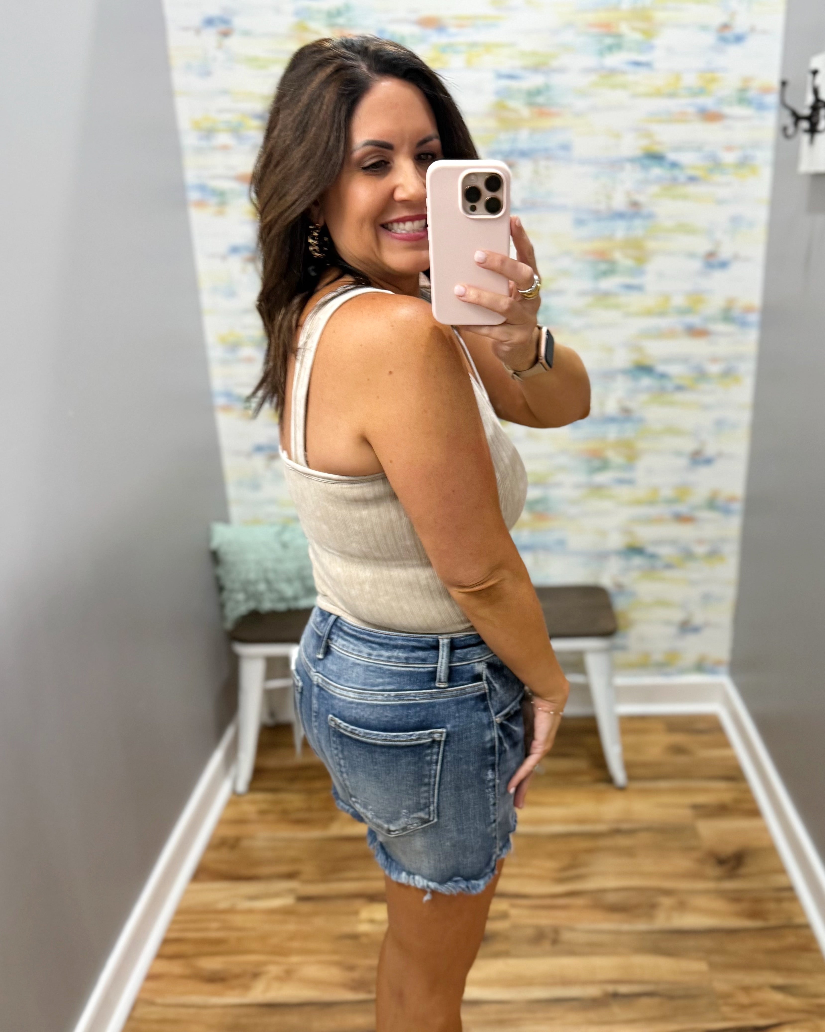 Woman taking a mirror selfie wearing a white tank top and denim shorts in a room with colorful wallpaper.
