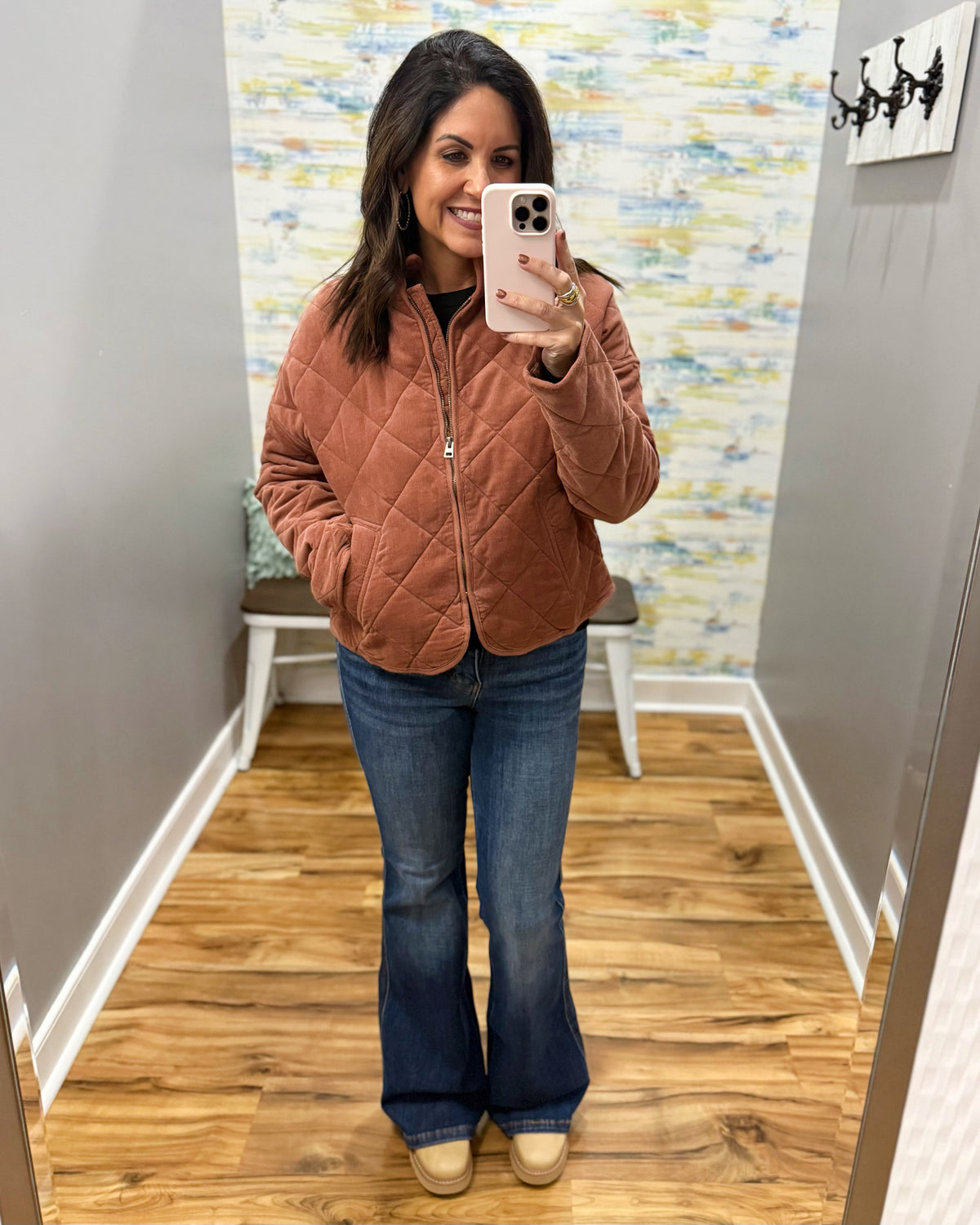 A woman takes a mirror selfie wearing a quilted rust-brown zip-up jacket, blue flare jeans, and beige shoes in a fitting room with wood floors and a colorful abstract wall.