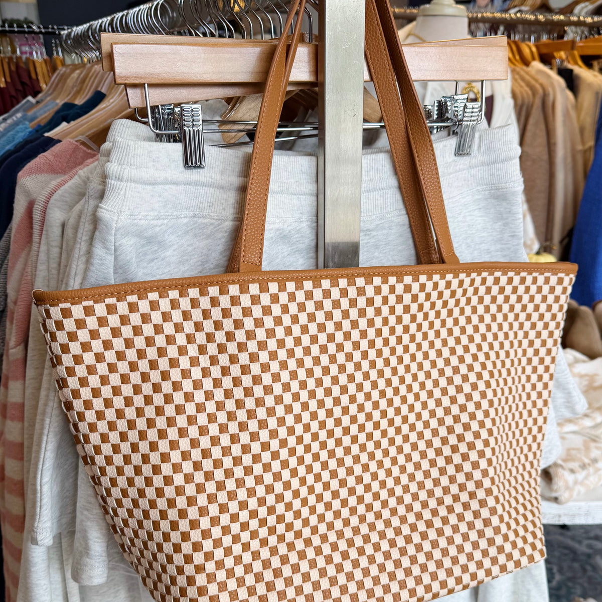 A large woven tote bag with a tan and cream checkerboard pattern and brown shoulder straps, hanging on a clothing rack in a retail store.