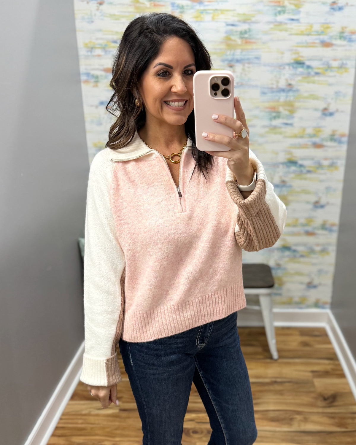A woman takes a mirror selfie wearing a light pink ribbed knit quarter-zip sweater vest over a cream long-sleeve top, paired with dark blue jeans.