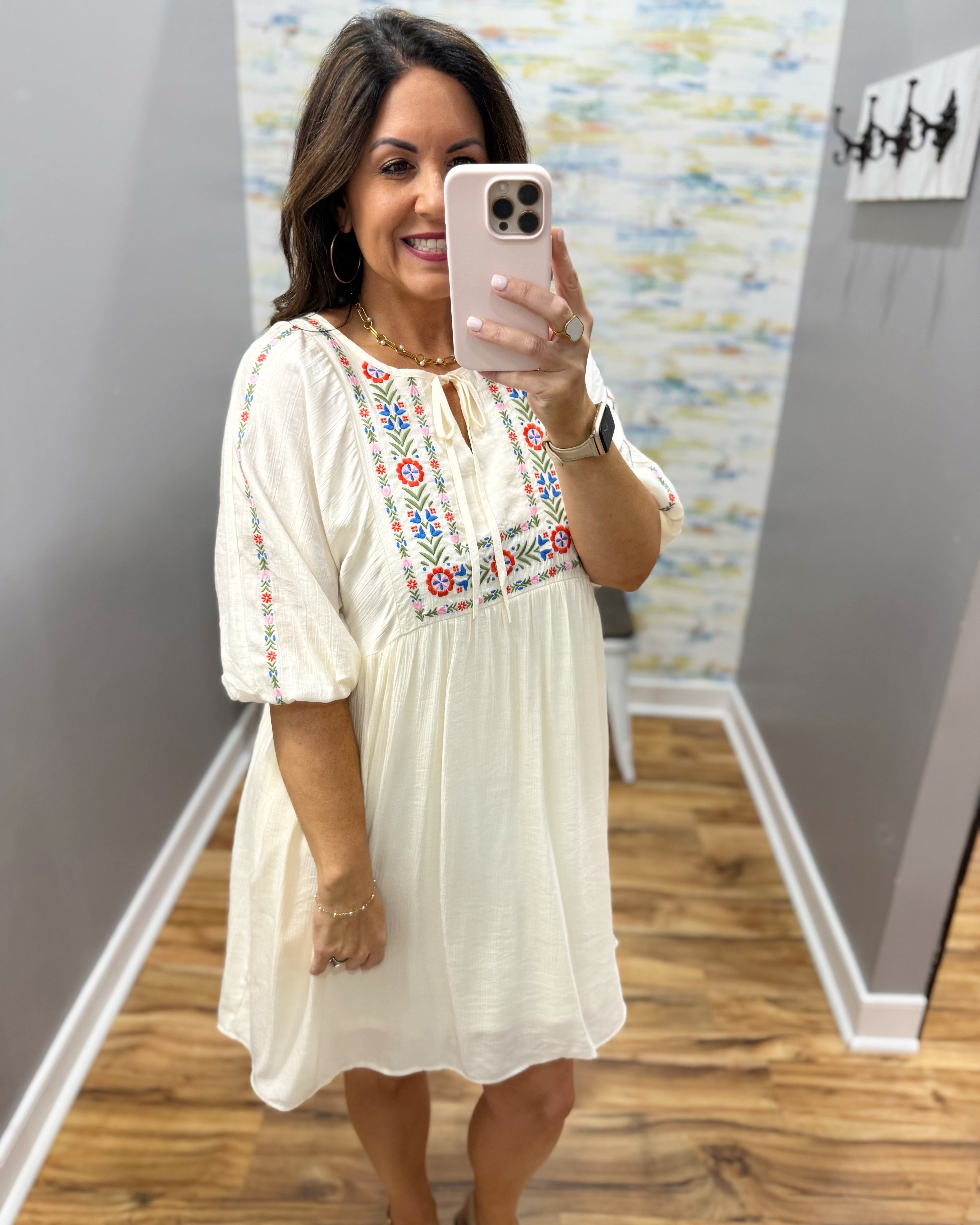 Woman taking a mirror selfie wearing a white dress with colorful embroidery in a room.