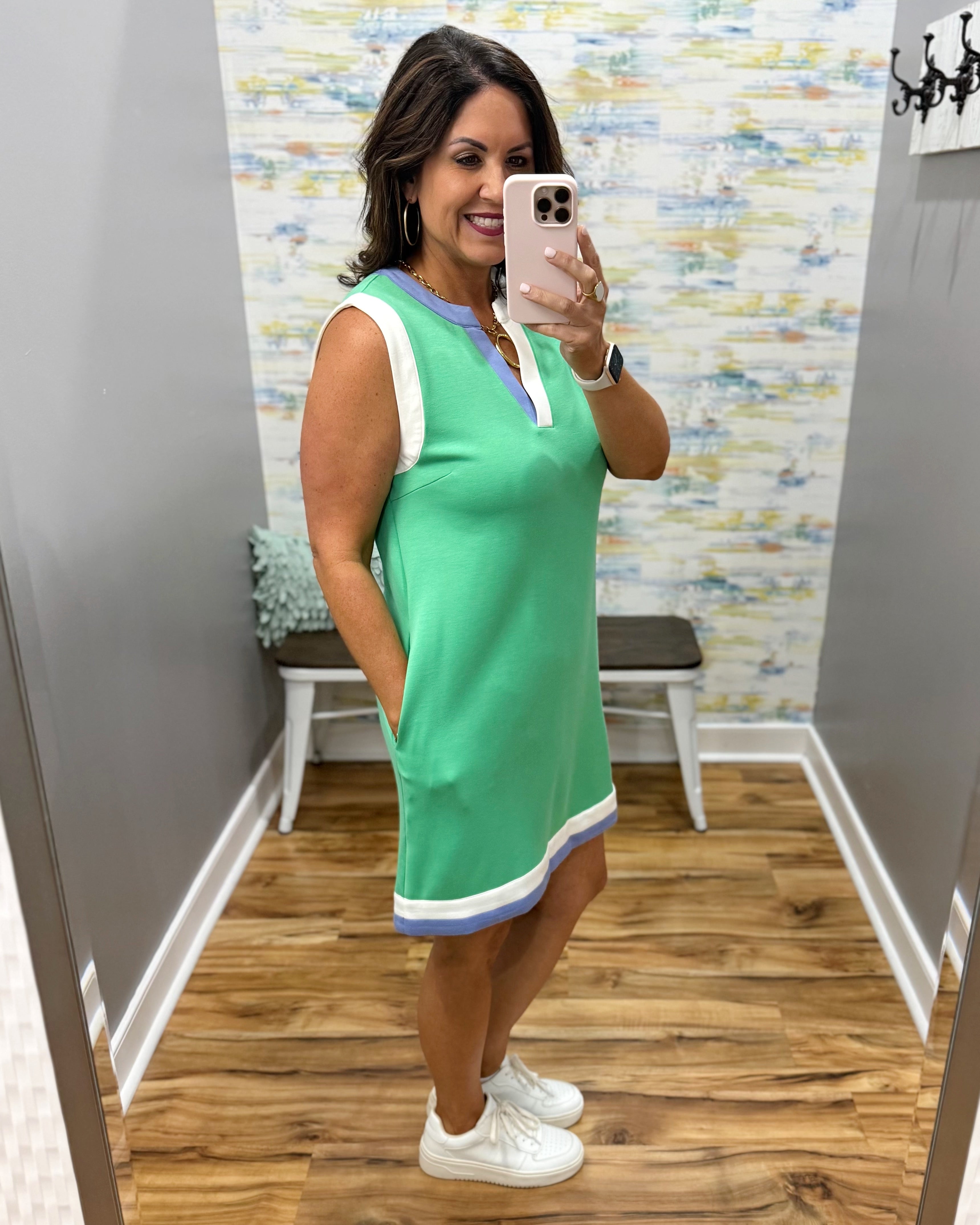 Woman taking a mirror selfie wearing a green dress with a colorful wall and wooden floor in the background.