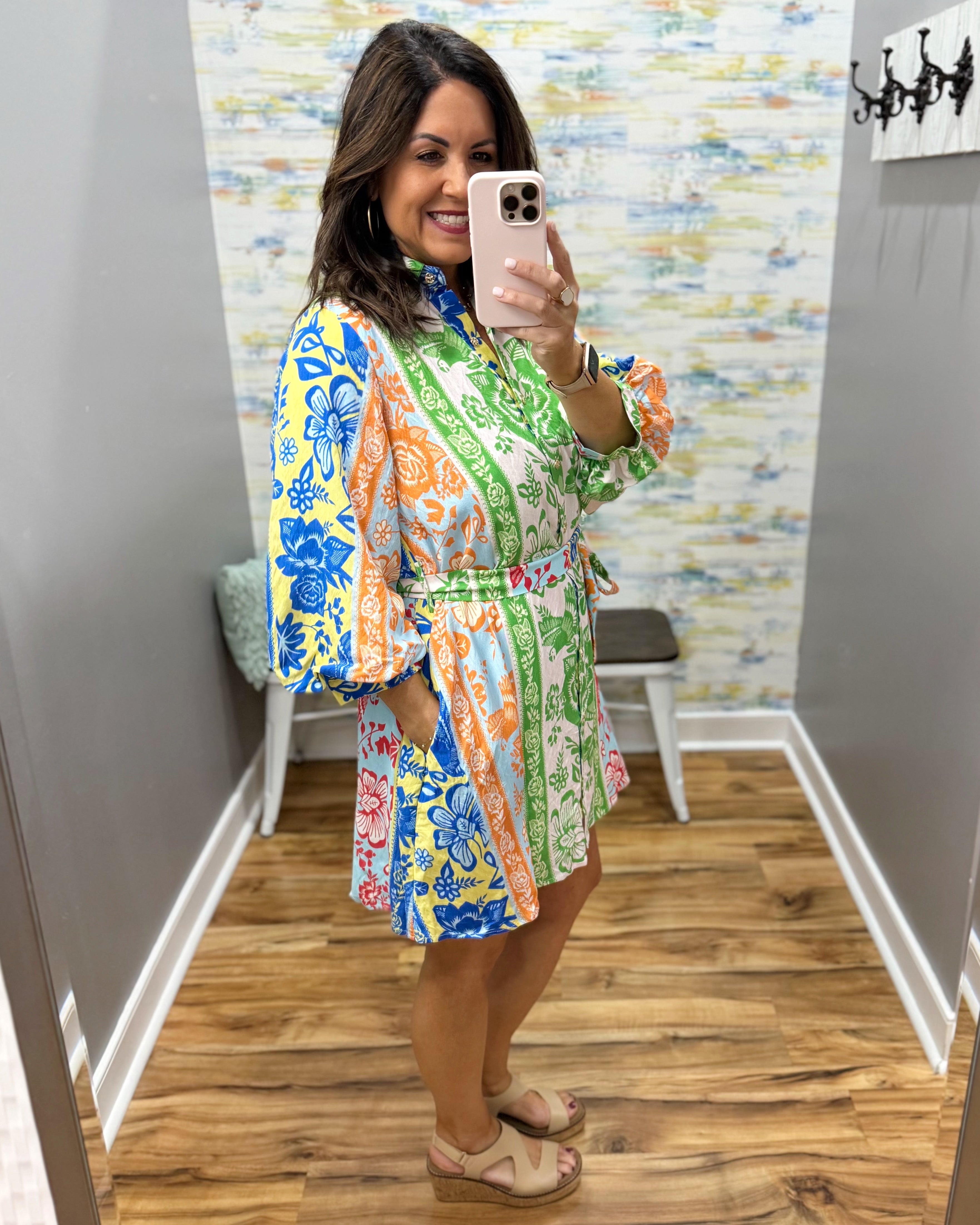 Woman taking a mirror selfie wearing a colorful floral dress in a room with a patterned wall.