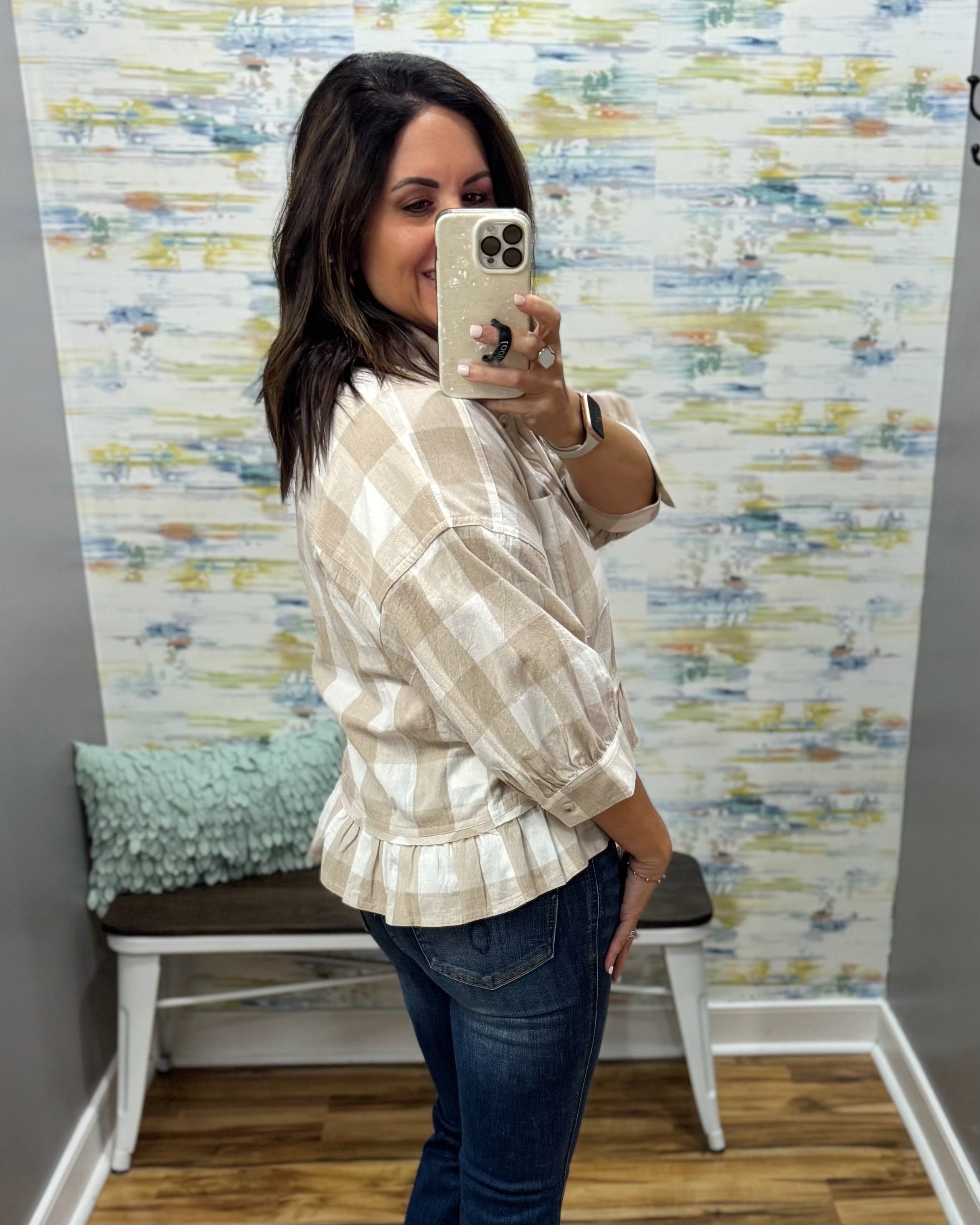 A woman takes a mirror selfie wearing a beige and white checkered peplum blouse with dark blue jeans, standing in front of a patterned wallpapered wall.