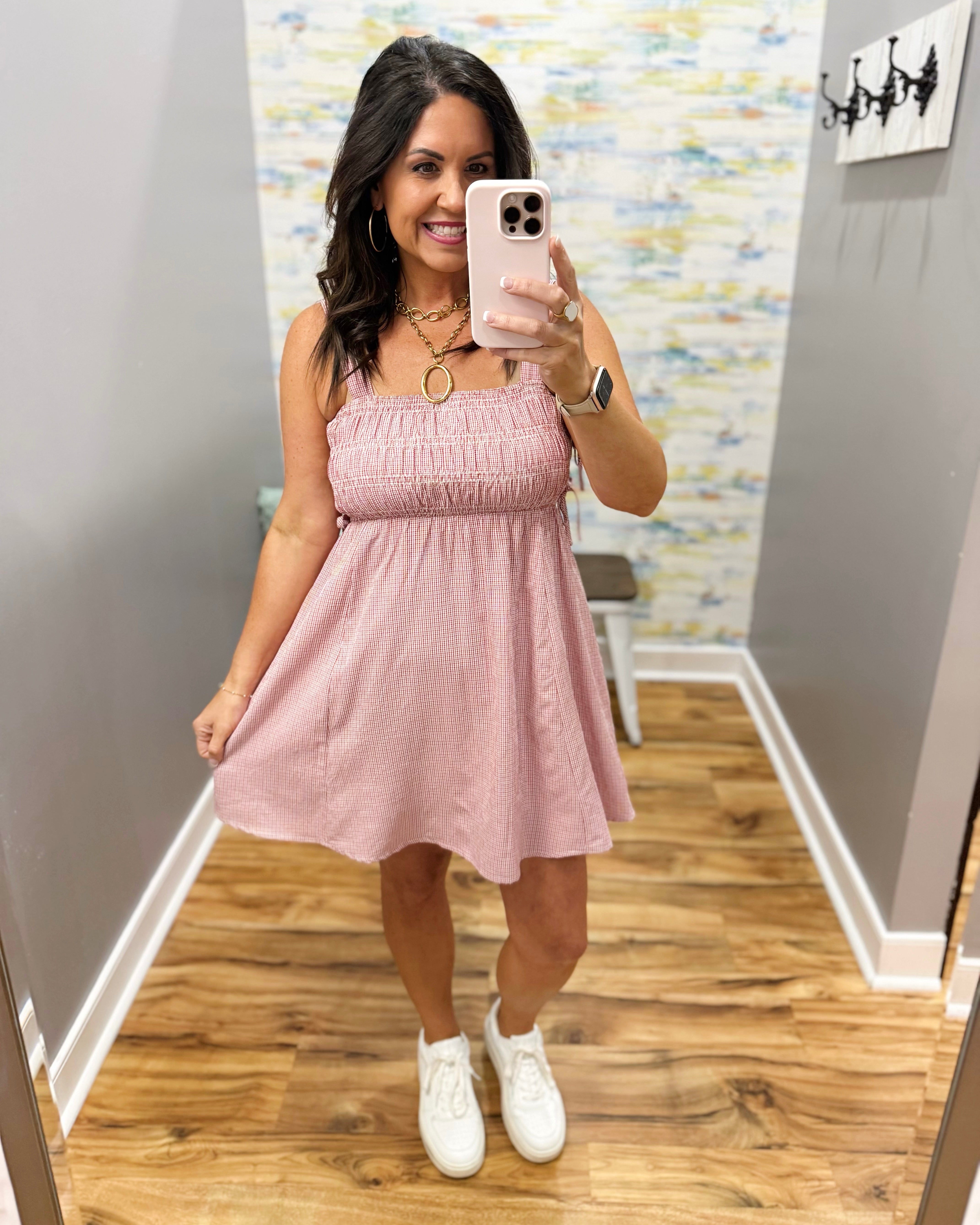 Woman taking a mirror selfie wearing a pink dress and white sneakers in a room with a colorful wall.